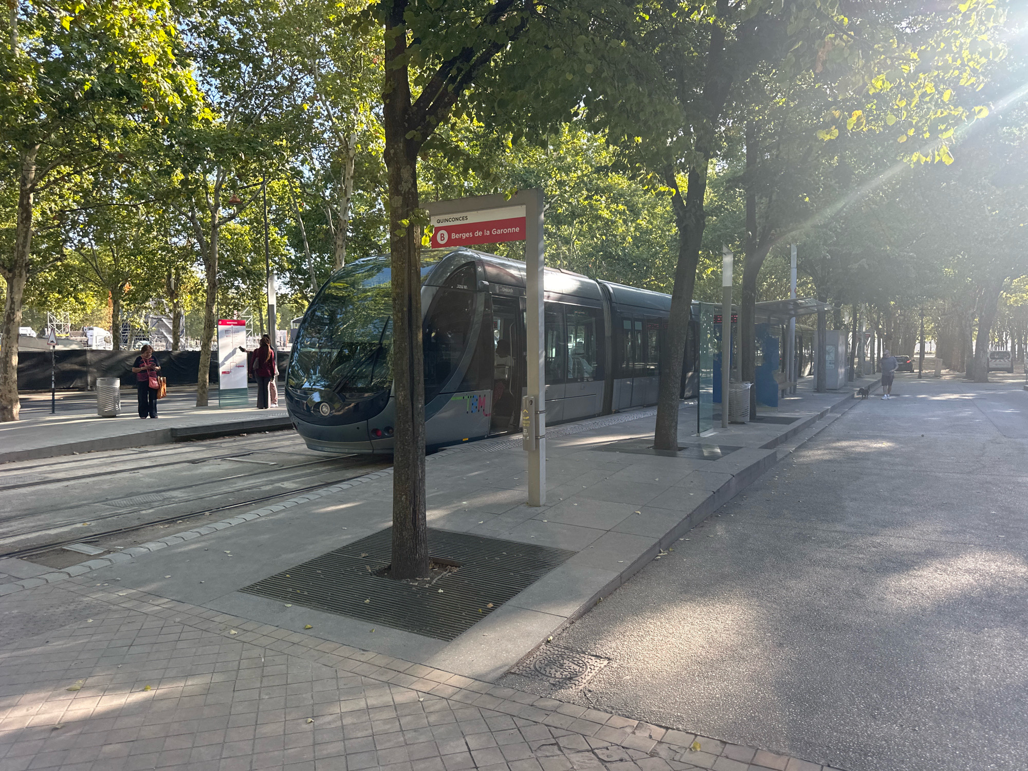The image depicts a sunny day scene at a tram stop in Bordeaux, France. In the foreground, a modern grey tram is stopped at a platform. The tram is articulated and has tinted windows. A red and white sign above the tram reads "QUINCONCES" and "B Berges de la Garonne," indicating this is the Berges de la Garonne stop on line B.
The platform is paved with light-colored square tiles and bordered by a grey concrete curb. Several trees with lush green foliage are planted along the platform, casting dappled shadows on the ground. One tree in the foreground has a metal grate surrounding its base.
On the platform, two individuals are visible. One is a woman standing with her back to the camera, wearing dark clothing and carrying a light-colored bag over her shoulder. Another person, also wearing dark clothing, is standing further down the platform, appearing to be looking at their phone. In the distance, other people and a dog can be seen walking along the paved road adjacent to the tram line.
The environment appears to be a wide avenue lined with mature trees. The sun is bright, creating lens flares and highlighting the leaves of the trees. The overall impression is that of a well-maintained urban public transportation hub on a pleasant day.