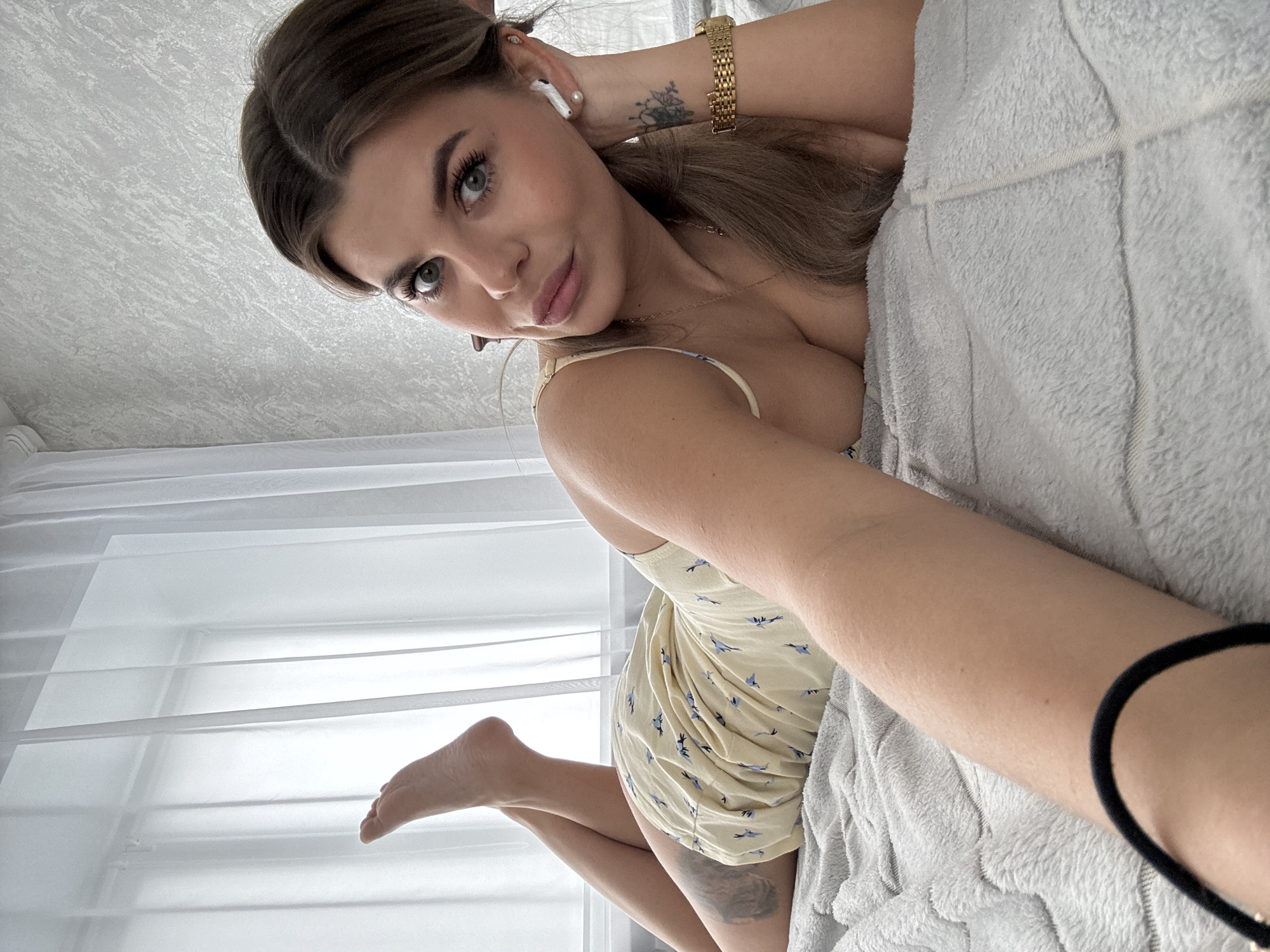 A woman is lying on a bed. She is wearing a light yellow dress with blue bird prints. She has a gold watch on her left wrist and a black hair tie around her right wrist. The background includes a window with white curtains and a textured white ceiling. The location is assumed to be Sarapul, Russia, based on the user's query context.