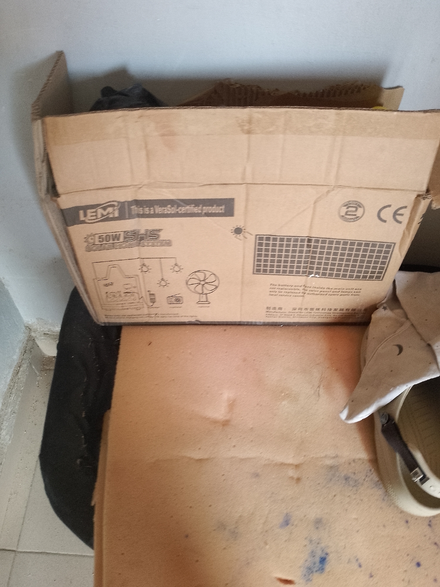 In Jauro Yinu, Nigeria, within an indoor environment, a cardboard box is positioned on top of a black object and a beige surface. The cardboard box displays text and graphics, including "LEMI," "This is a Verasol-certified product," "50W SHS SOLAR HOME SYSTEM," a diagram of a solar home system with components like a main unit, lamps, a fan, and a radio, a solar panel graphic, a circular logo with the number "2," and the letters "CE." A portion of a tan-colored shoe or sandal is visible on the right side of the image, resting on the beige surface. The background consists of a light gray wall. The lighting is ambient, with no specific light sources clearly identifiable.