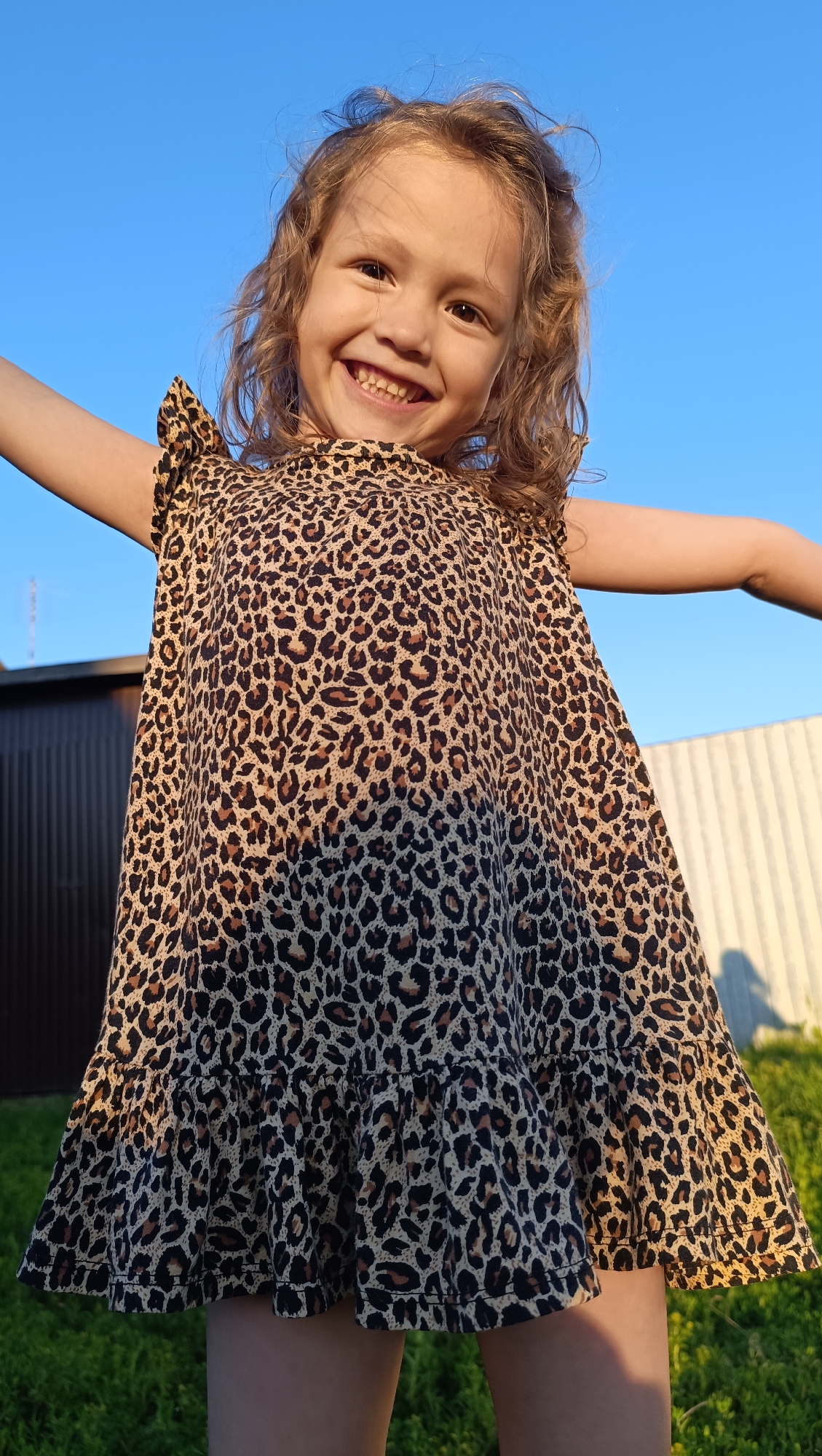 In the city of Karsha, Russia, a young Caucasian girl with curly blonde hair is captured in a low-angle shot, smiling broadly at the camera. She is wearing a leopard-print dress with ruffled sleeves and a tiered hem. Her arms are outstretched, and she appears to be mid-motion, perhaps jumping or dancing. The background consists of a clear blue sky, a dark-colored shed or fence on the left, and a light-colored fence with vertical slats on the right. A patch of green grass is visible at the bottom, illuminated by what appears to be late afternoon or early evening sunlight, casting a shadow on the ground. The image is brightly lit, highlighting the girl's joyful expression and the details of her dress.