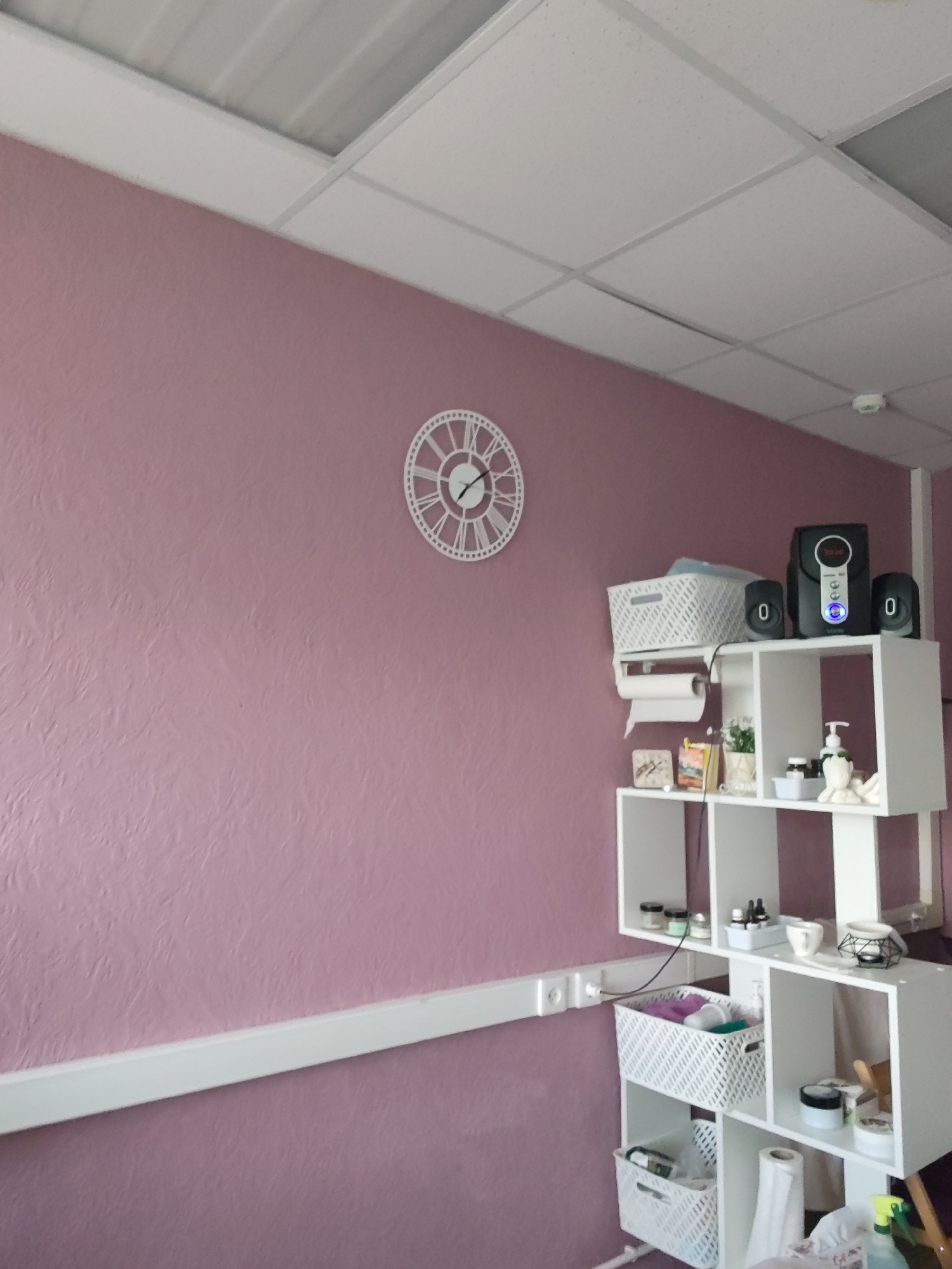 The image shows an interior scene. A pink wall has a white decorative clock on it. Beneath the clock, a white shelf unit holds a variety of items, including a basket, speakers, and toiletries. A white molding runs along the wall near the bottom of the frame. The setting appears to be indoors, likely a room in a commercial or residential building. The photograph may have been taken in Chaikovsky, Russia, as requested, but there is no visual confirmation of this location in the image.