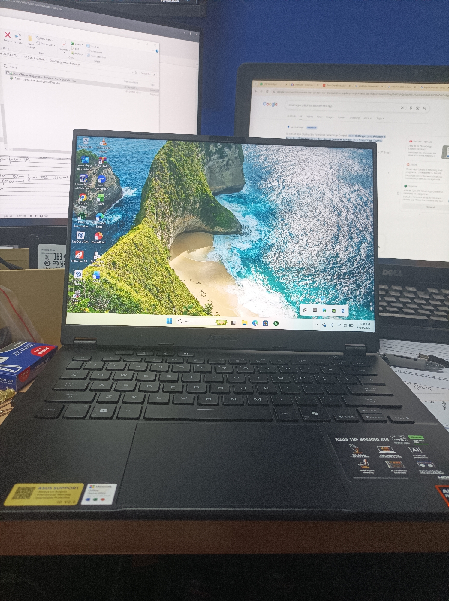 A laptop with a scenic wallpaper of a cliffside beach is open on a desk. The laptop screen displays several application icons including Google Chrome, Excel, and Microsoft Edge. A second monitor is visible behind the laptop, displaying a file explorer window. A keyboard and desk accessories are also present. The scene appears to be in an office or home setting. The location of Sarajaya, Indonesia is not evident in the image.