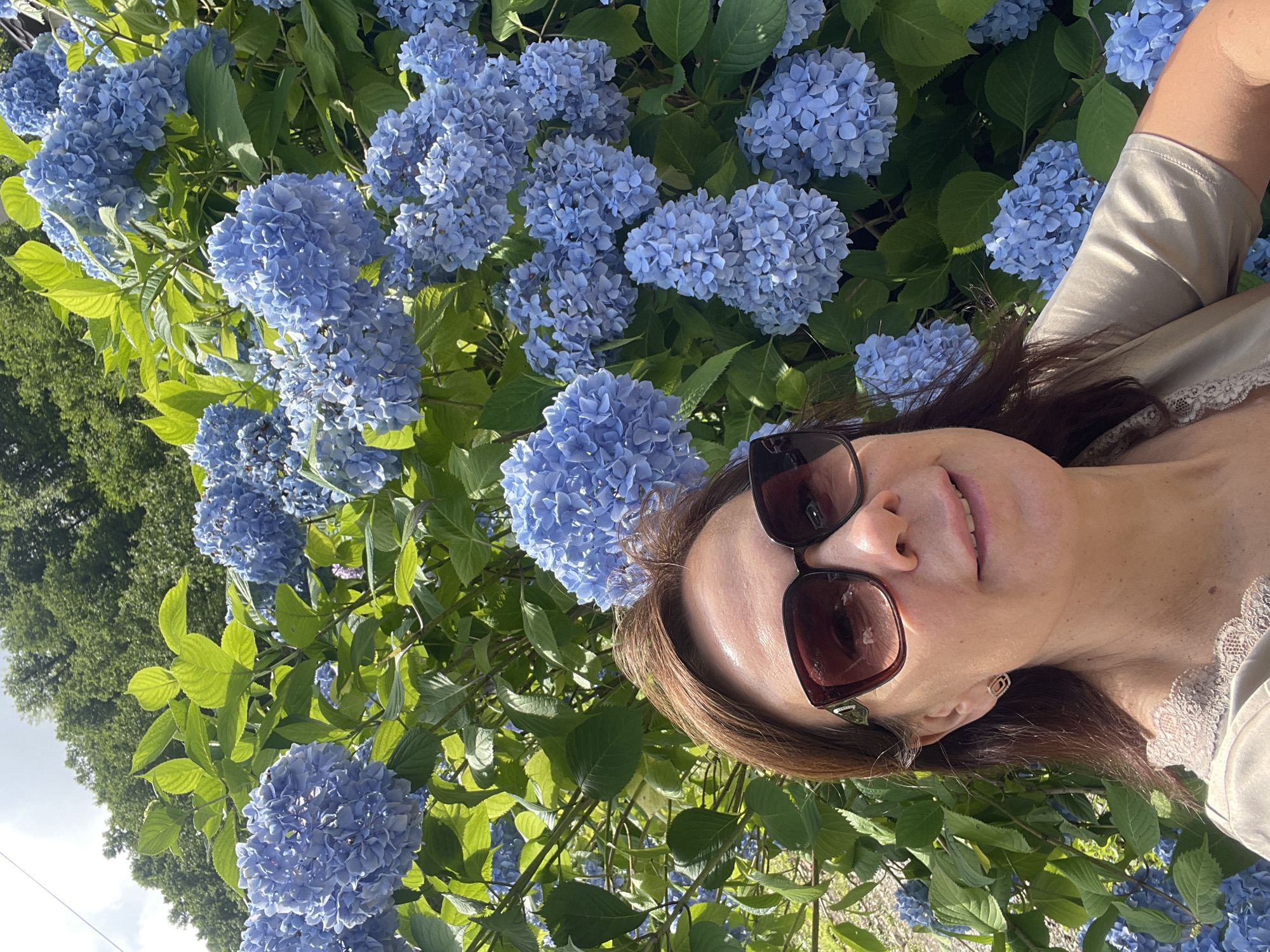 A selfie taken by a woman outdoors in Tqwarchal, Abkhazia, shows her smiling in front of a large hydrangea bush in full bloom. The woman has brown hair, is wearing large, dark-rimmed sunglasses, and a light-colored top with lace trim. The hydrangea flowers are a vibrant blue, and the leaves are a lush green. The background features trees and a partly cloudy sky. The scene suggests a sunny day in a natural setting.