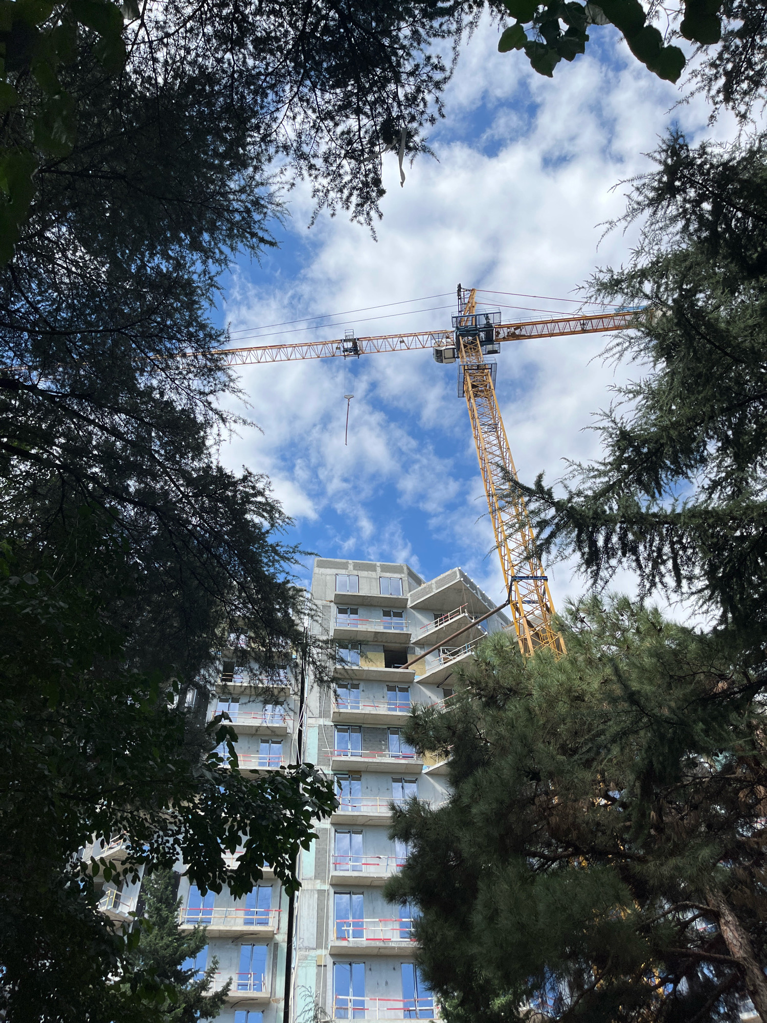 An outdoor, upward-facing, eye-level shot captures a construction site in Tbilisi, Georgia, framed by dense, dark green trees. The sky is bright blue with scattered white clouds. A tall yellow construction crane dominates the upper half of the frame, its long arm extended horizontally. Below the crane, a multi-story building under construction is visible, its facade partially covered in grey material and featuring multiple floors with windows. Balconies with red and white safety railings are present on several levels. Some windows are already installed, reflecting the sky. The building appears to be in the early to middle stages of completion. The surrounding trees create a natural frame, with some leaves and branches obscuring parts of the view, adding a sense of depth and contrast to the urban development. No people are visible in the image.