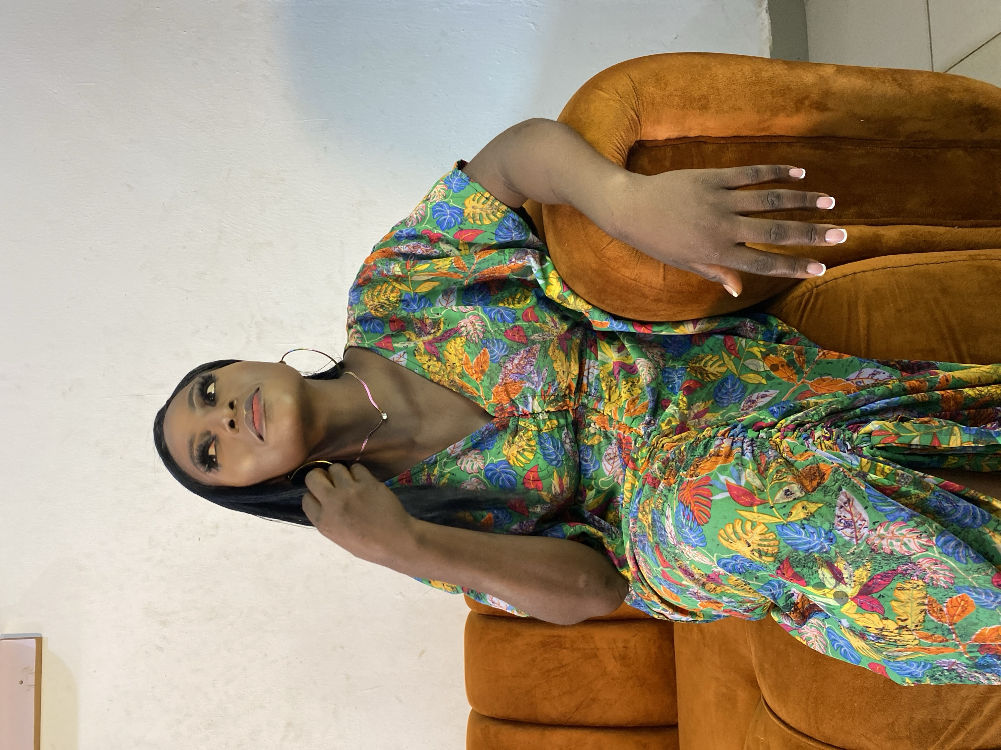A young Black woman is seated on an orange velvet sofa, looking upwards and to the right. She has long, dark hair, is wearing a brightly colored, floral-patterned dress, and hoop earrings. Her left arm is bent, with her hand resting on her lap, and her right arm is resting on the back of the sofa. The background is a plain, textured white wall.
