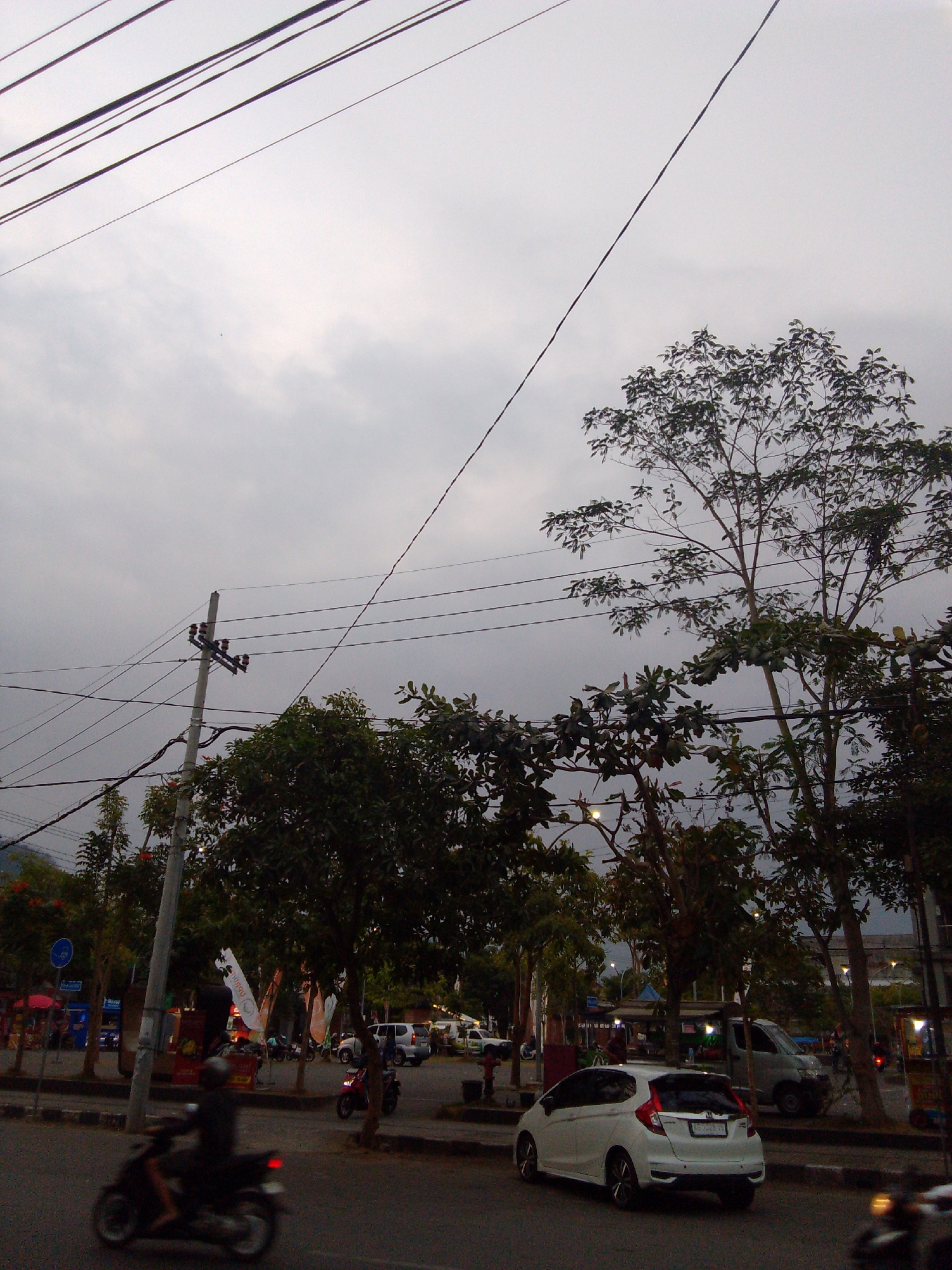 The image depicts a street scene in Trenggalek, Indonesia, likely during overcast daytime or twilight. Prominent overhead power lines dominate the upper portion of the frame. In the mid-ground, a street is visible with several vehicles, including a white hatchback car in the foreground, a light-colored van, and motorcycles. Trees line the street, some with sparse foliage. A utility pole with electrical insulators stands on the left side of the road. Further back, buildings and market stalls are partially visible. On the right side of the street, a small red motorcycle is parked. A lone figure is riding a dark-colored motorcycle in the foreground, moving away from the camera. The overall atmosphere is urban and slightly gloomy due to the weather conditions.