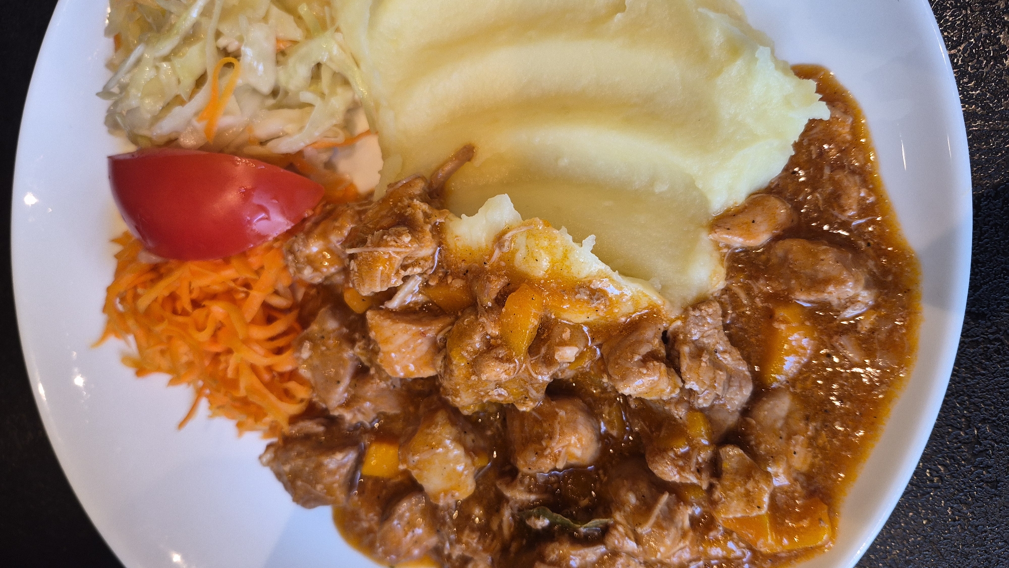 A close-up, overhead shot shows a white plate filled with a meal. On the left is a portion of stewed meat in a thick, brown sauce with visible pieces of carrot. To the right of the stew is a mound of mashed potatoes. At the top of the plate, from left to right, are shredded carrots, a wedge of tomato, and a small serving of coleslaw. The plate rests on a dark, textured surface. The image is a food photograph, and there is no indication of a specific location in Daugavpils, Latvia.
