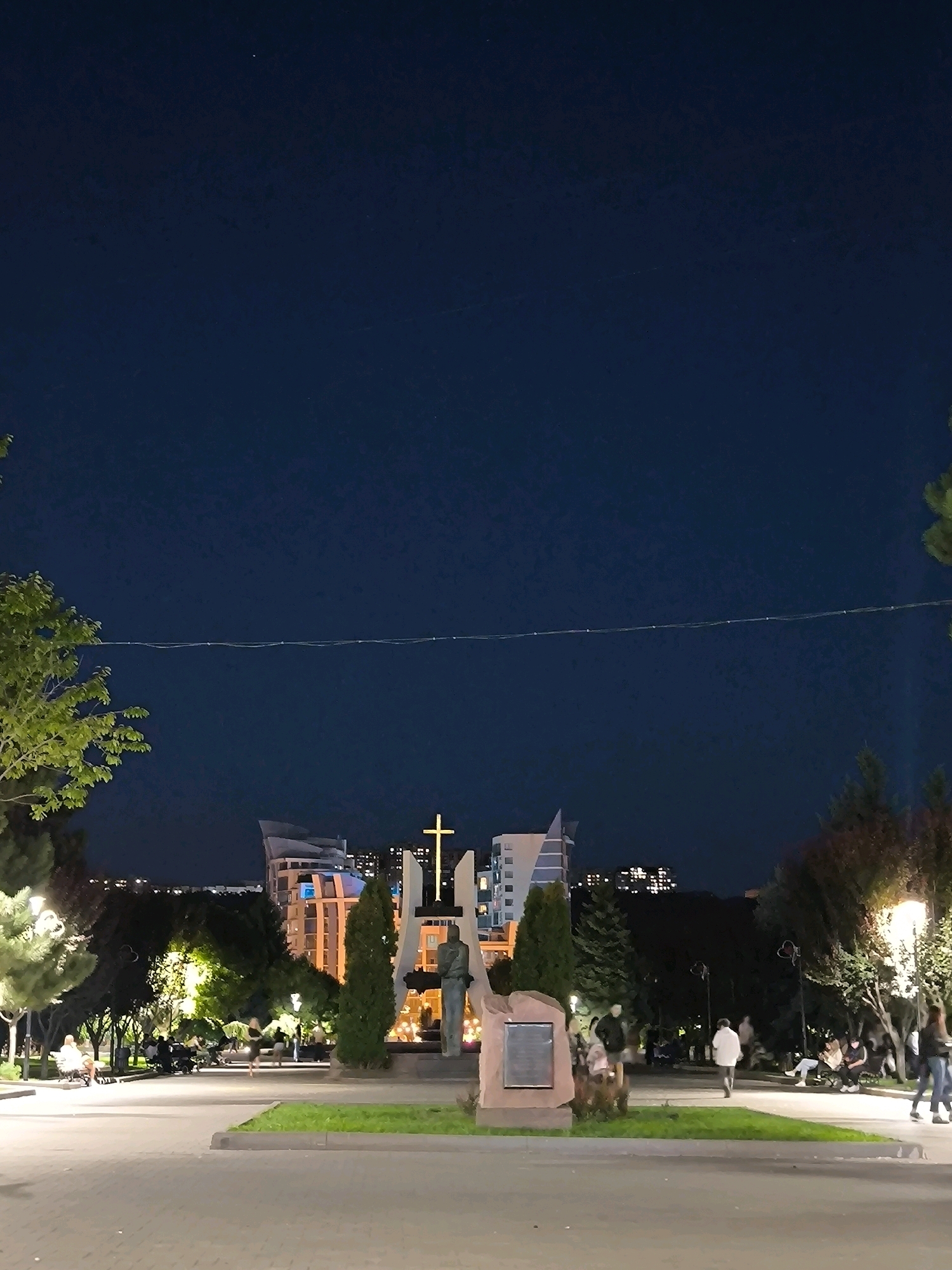 The image depicts a nighttime view of a public square in Chișinău, Moldova. In the foreground, a paved walkway leads towards a monument featuring a statue and a large cross illuminated by lights. The monument appears to be made of stone and possibly metal. Several trees are scattered throughout the scene, some with decorative lighting. In the background, modern buildings with illuminated windows are visible against the dark night sky. People are seen walking and sitting on benches in the square, enjoying the evening. Streetlights cast a warm glow on the pathways and surrounding areas. A thin wire, possibly a string of lights, stretches across the sky in the middle distance. The overall atmosphere is calm and serene, suggesting a leisurely evening in a well-lit urban park.