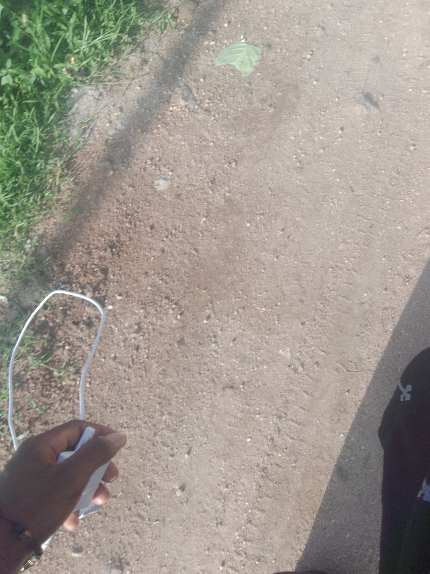 The image shows a hand holding a white USB charger in Jalingo, Nigeria. The hand, adorned with beaded bracelets, is positioned in the lower left of the frame. The charger is white and appears to be unplugged. In the background, a patch of green grass borders a dirt path. The lighting suggests it is daytime, with shadows cast on the path. A dark shadow partially covers the right side of the image, obscuring the lower right corner.
