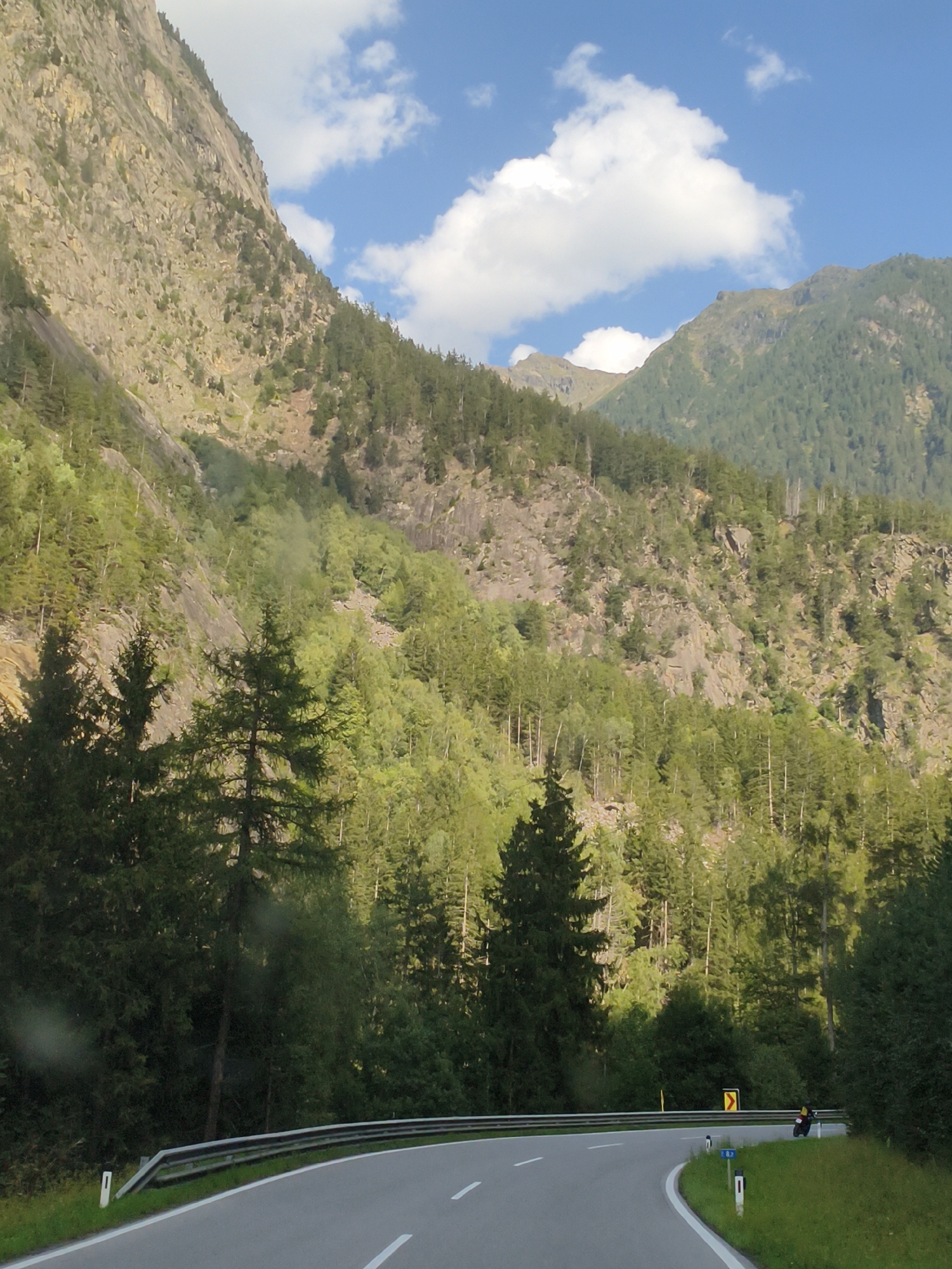 The image captures a scenic view of a winding mountain road in Tumpen, Austria, on a clear day. The road is paved and has white lane markings. On the left side of the road, a metal guardrail runs along the edge, separating it from the steep, rocky mountain face. The mountain is covered with dense green vegetation, primarily pine trees, that extend upwards towards the summit.
In the foreground, the road curves to the right. A motorcyclist, wearing a yellow jacket and a black helmet, is visible on the right side of the road, leaning into the curve. The motorcyclist appears to be alone and is in motion.
To the right of the road, there is a grassy embankment with a yellow directional sign pointing to the right, indicating a curve in the road. Several small blue and white marker posts are also visible along the roadside.
The background is dominated by the imposing mountain range, with its rugged slopes and a thick forest of trees. The sky above is a vibrant blue, dotted with fluffy white clouds, suggesting fair weather. The overall impression is one of a beautiful, natural landscape and an open road, inviting exploration.
