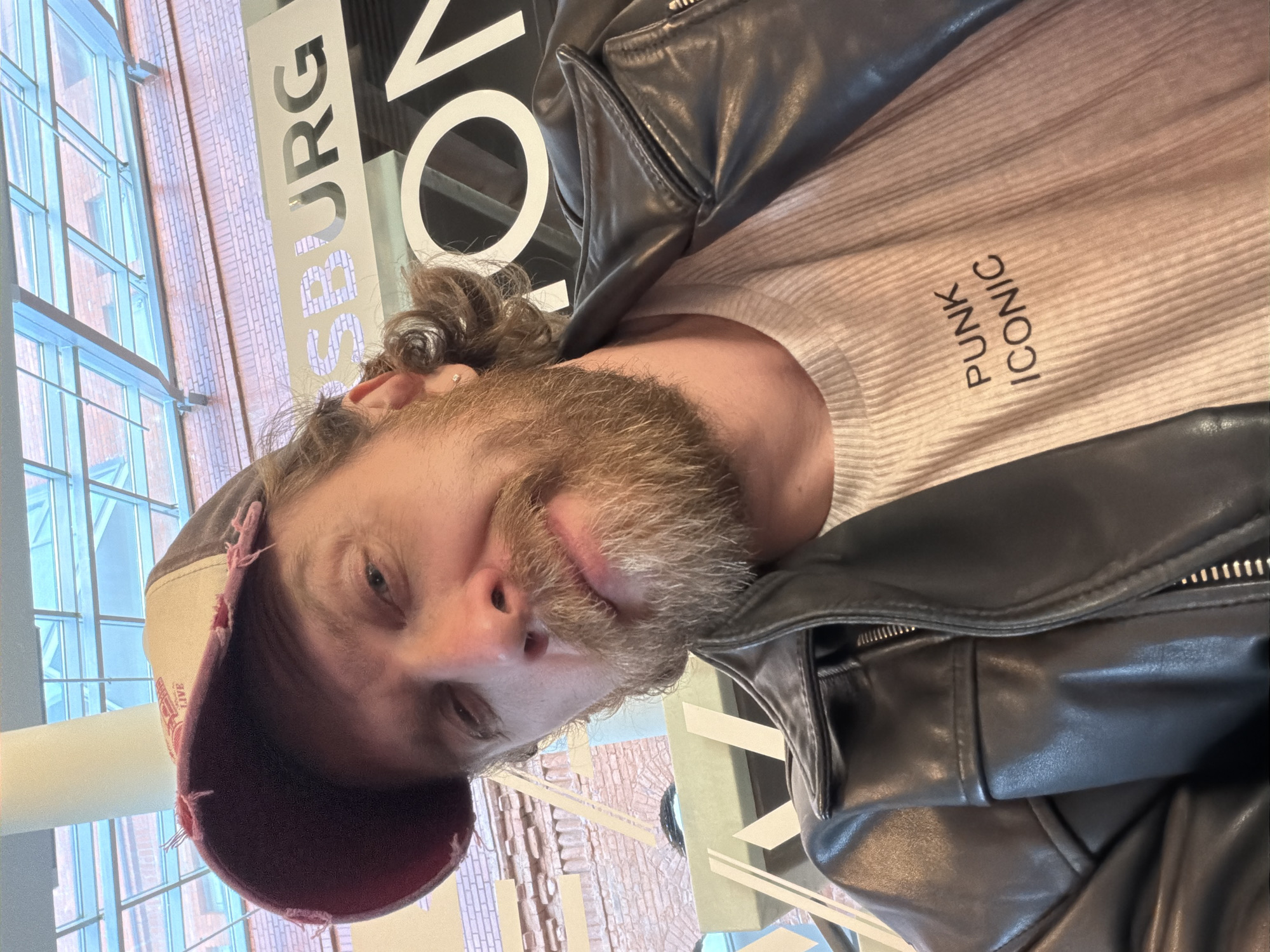 A medium close-up, slightly tilted portrait shows a man with a light brown beard and curly hair, wearing a black leather jacket, a light beige ribbed long-sleeved shirt, and a distressed burgundy and beige baseball cap. He is looking directly at the camera with a subtle, calm expression. The cap is worn backward, revealing its tattered edges.
The shirt features vertical text in black, reading "PUNK" above "ICONIC." The black leather jacket has visible zippers and seams, suggesting a modern or biker-style design.
The setting appears to be indoors, likely a modern building, with large, multi-paned windows in the background. Through these windows, an exterior brick building is visible, hinting at an urban environment. A large, prominent sign with white lettering on a black background is partially visible behind the man's head and shoulder. The discernible parts of the sign read "SBURG" and "ON," strongly suggesting a reference to Saint Petersburg given the provided location context. Natural daylight illuminates the scene, indicating it is daytime. The man appears to be taking a selfie or being photographed casually.