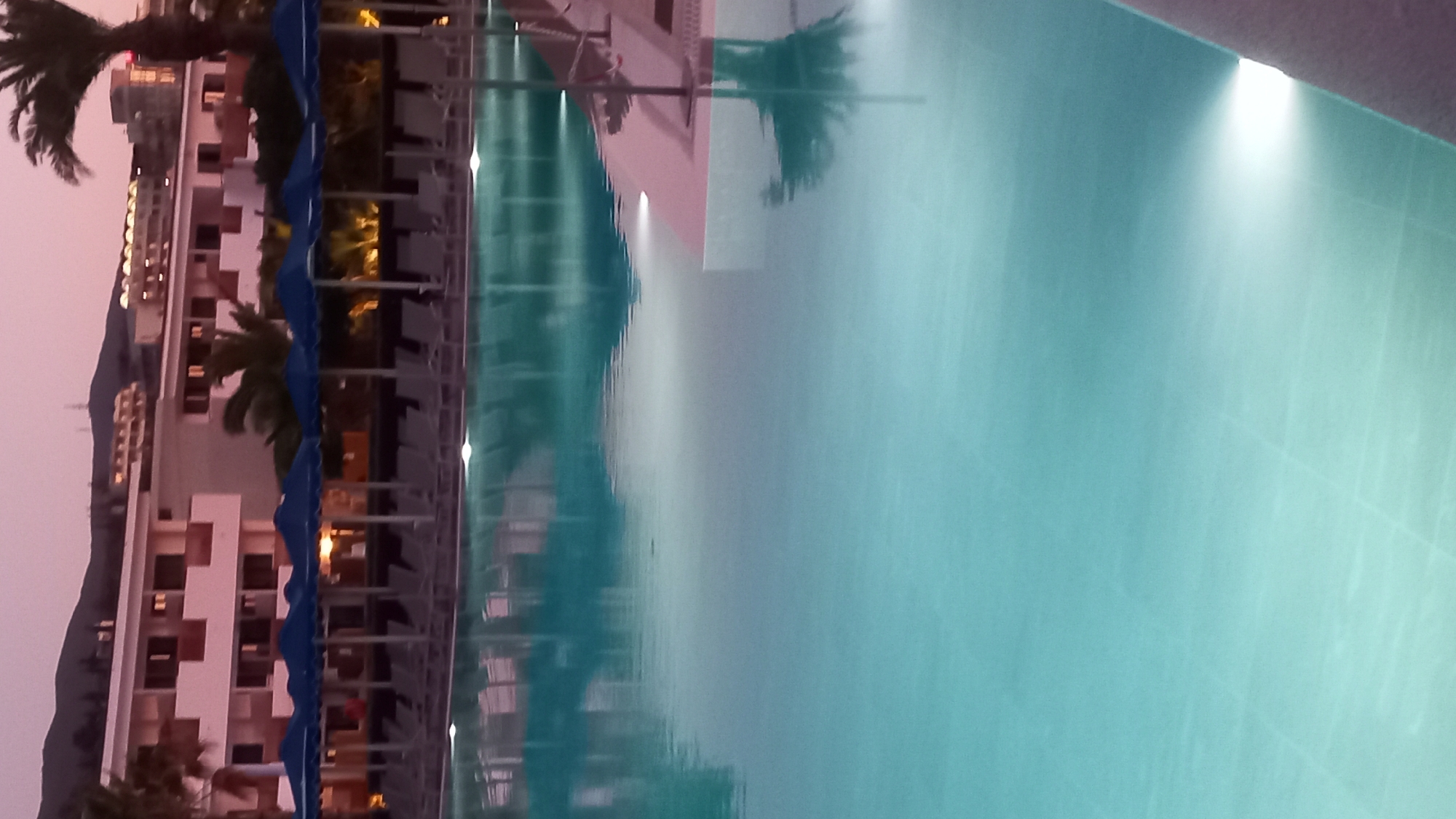 The image shows a swimming pool at dusk. The pool water is a bright turquoise color and reflects the lights from the hotel buildings and the sky. In the background, there are several hotel buildings and some palm trees. The sky is a soft pink and purple color, indicating the end of the day. The scene appears to be a resort or hotel complex, likely located in Kos, Greece, given the architectural style and vegetation often found on Greek islands.