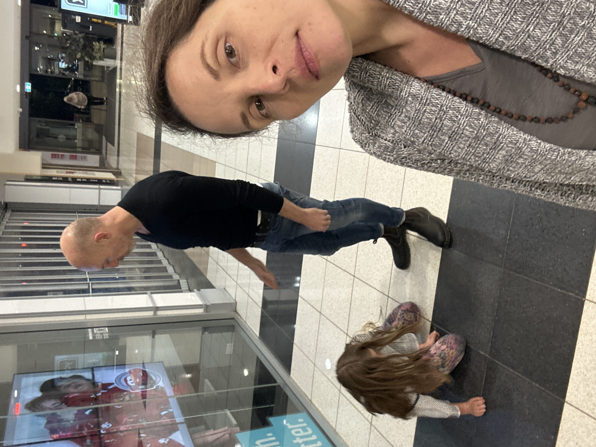In Toormina, Australia, within a shopping center hallway, a bald man in a black long-sleeved shirt and blue jeans stands looking at a large display screen. To his left, a young girl with long brown hair is kneeling on the floor, seemingly playing or examining something. In the foreground, a woman with brown hair and a grey patterned cardigan is taking a selfie, her face angled towards the camera. The hallway features a tiled floor with contrasting black and light-colored sections, glass storefronts, and automatic doors in the background leading to an exit.