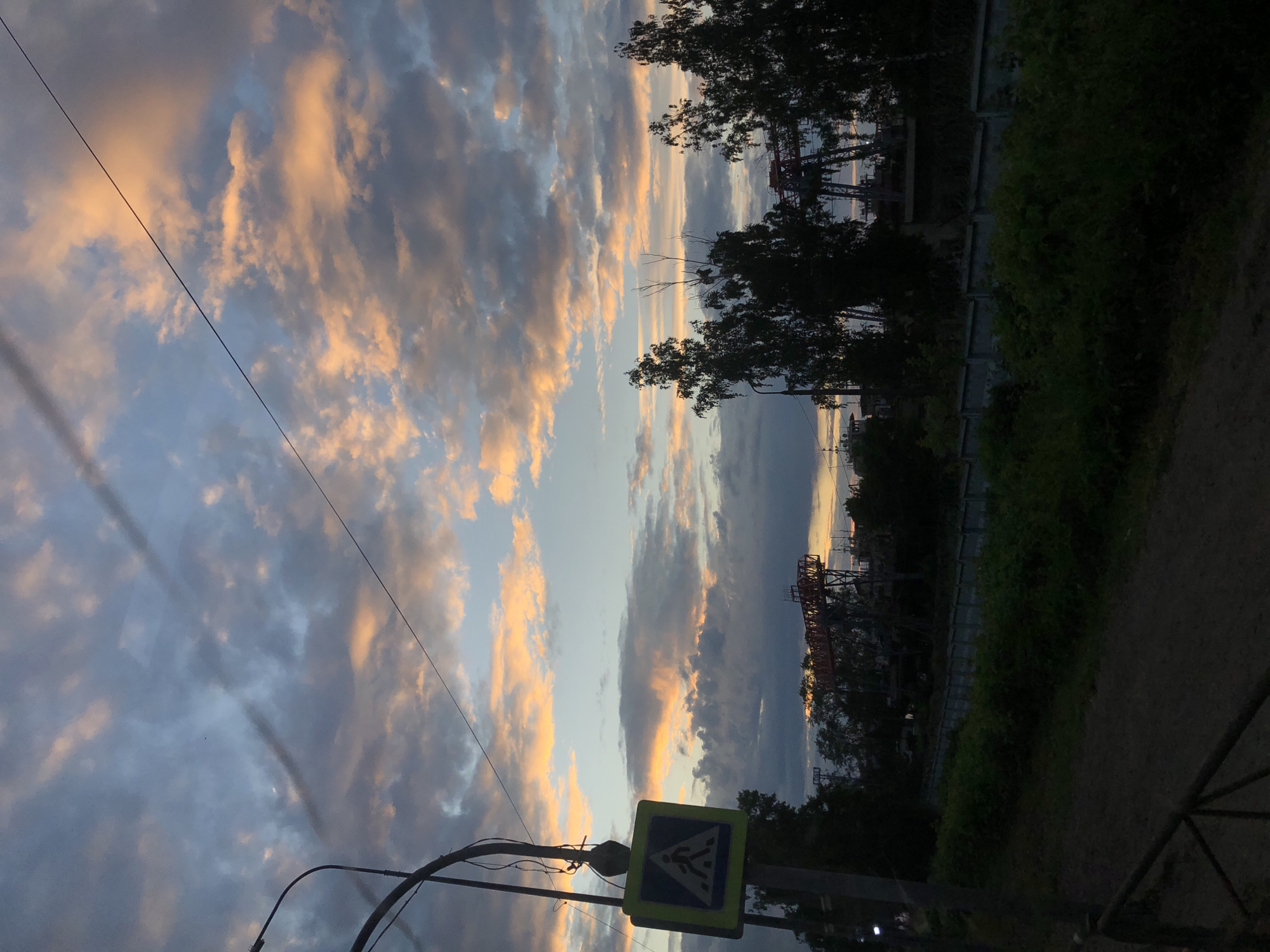 The image depicts a street scene in Chaikovsky, Russia, during twilight. A pedestrian crossing sign is prominently displayed on a lamppost in the foreground. Beyond the sign, trees line a concrete fence that separates the street from a more industrial area. In the distance, the silhouettes of industrial cranes are visible against a sky filled with dramatic clouds tinged with orange and pink from the setting sun. The overall atmosphere is one of an urban landscape transitioning into night.