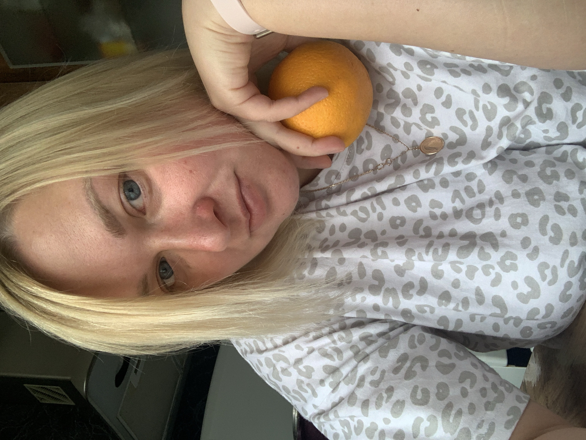 A selfie-style portrait, rotated 90 degrees counter-clockwise, captures an individual with blonde hair and blue eyes in an indoor setting in Chaikovsky, Russia. The light-skinned person looks directly at the camera with a neutral expression, their blue eyes prominently visible. They are wearing a light-colored garment, likely a t-shirt, featuring a gray leopard print pattern. A delicate gold-colored chain with a small, round pendant hangs around their neck. On their right wrist, a light pink or beige smartwatch or fitness tracker is visible. The individual's right hand is holding a whole, unpeeled orange, positioned close to their face. The background is dark and blurred, offering no specific location cues beyond an interior space. The time of day and weather are indeterminate. No visible text is present in the image.