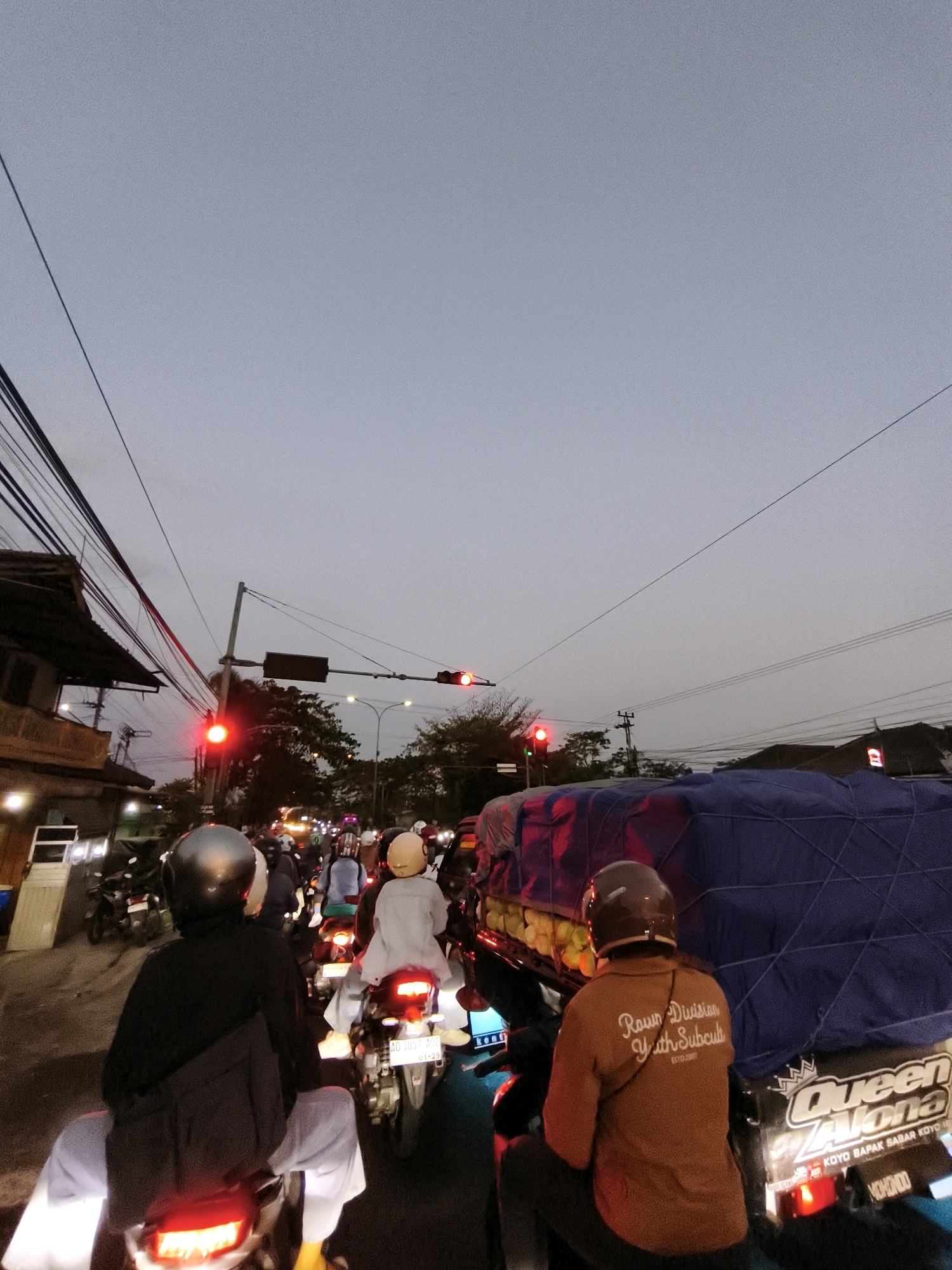 The image depicts a traffic jam on a street in Teras, Indonesia, during dusk or early evening. The sky is a pale, muted blue, indicating the fading light. Overhead, a tangle of electrical wires crisscrosses the frame, supported by poles. On the left side of the road, a two-story building with a dark, sloped roof and visible windows is present.
The street is filled with a dense congregation of motorcycles and a few other vehicles. Many people are riding on motorcycles, wearing helmets and casual clothing. Some riders are looking ahead, while others appear to be stationary in the traffic. Red traffic lights are visible at an intersection, contributing to the standstill.
In the foreground, the back of a motorcycle with a person wearing a dark helmet and a black jacket is prominent. To the right of this motorcycle, another rider in a brown shirt with white lettering, possibly a logo or text, is also on a motorcycle. This rider's motorcycle is carrying a large, blue-covered load, possibly goods, behind them, with visible yellow items peeking out from underneath. A large graphic with the word "Queen" and "Alona" is visible on the side of the vehicle carrying the load. In the background, more motorcycles and the lights of other vehicles are visible, creating a sense of depth and the extent of the traffic congestion. The overall scene captures a typical urban traffic situation in Indonesia at the end of the day.