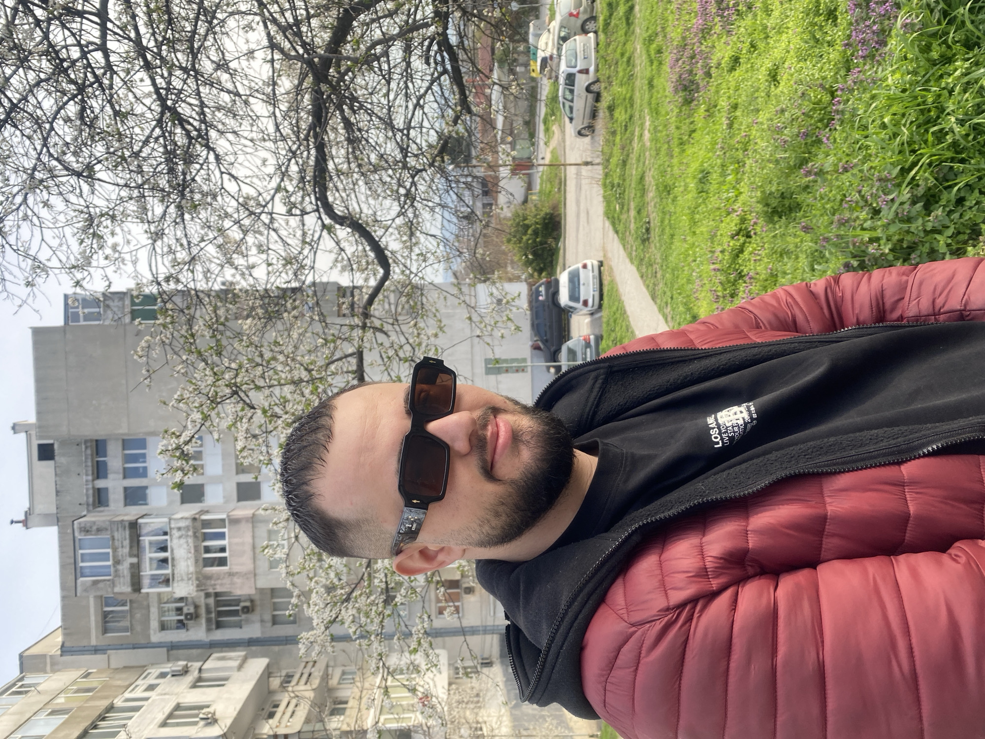 A man with dark hair and a beard is wearing sunglasses, a black hoodie, and a red puffer jacket. He is standing outdoors, and behind him is a residential building with many windows. A tree with white blossoms is partially visible, obscuring some of the building. In the background, there is a street with parked cars and a grassy area. The picture was taken in Varna, Bulgaria.