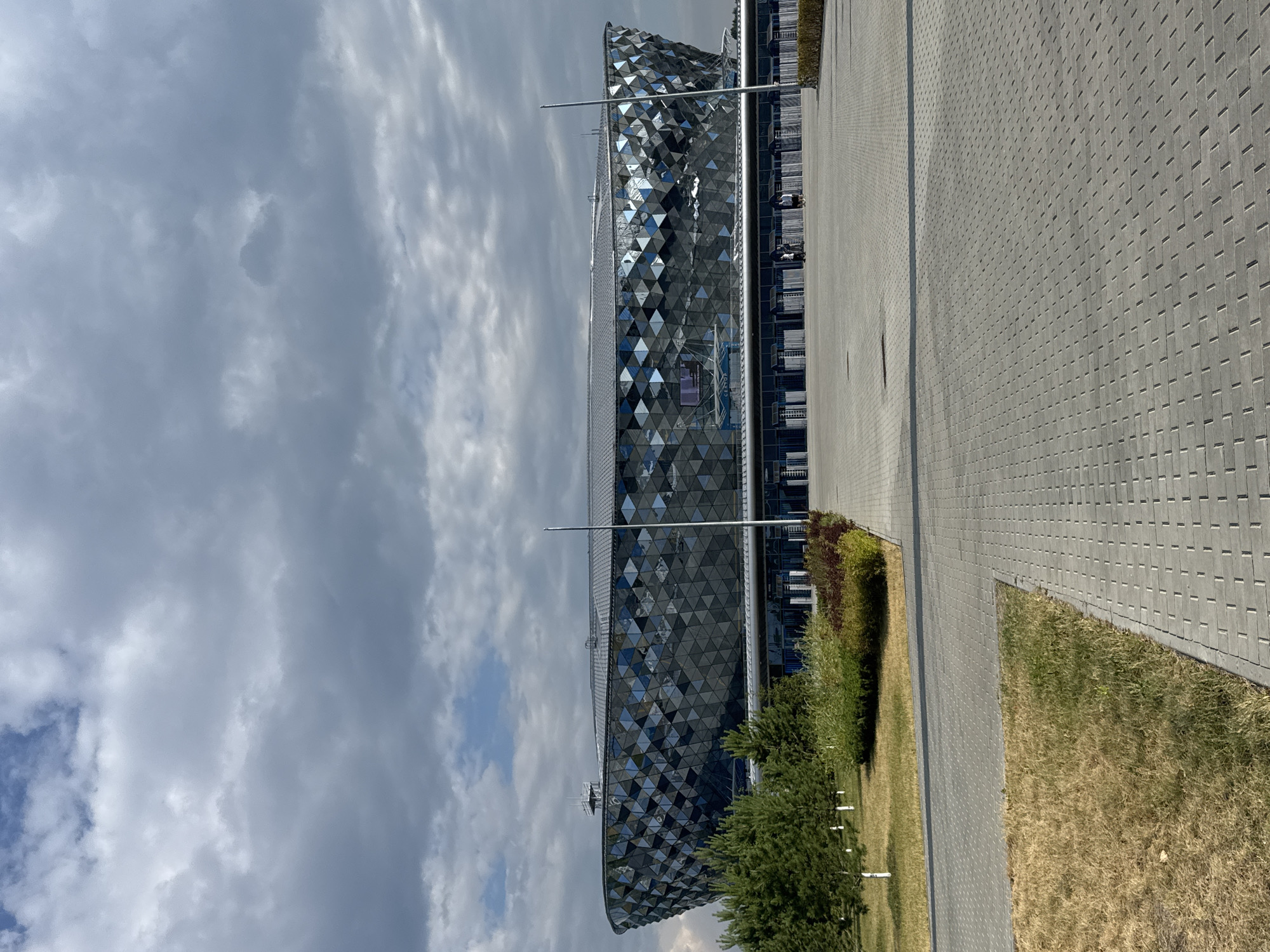 The image shows the exterior of the "Lokomotiv" Stadium in Novosibirsk, Russia. The stadium has a modern, faceted design with a reflective facade composed of triangular panels. The sky above is partly cloudy, with dramatic gray clouds interspersed with patches of blue sky. In the foreground, there is a paved walkway made of grey interlocking bricks, leading towards the stadium. To the left of the walkway, there is a patch of dry, yellowed grass and a landscaped area with green bushes and some trees. Several people are visible in the distance, walking near the entrance of the stadium. The overall scene depicts a large, contemporary sports venue under a dynamic sky.