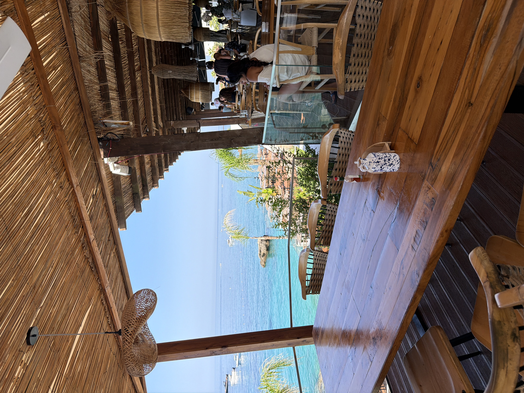 A wide shot captures a seaside restaurant in Himarë, Albania, with a focus on a large, polished wooden table in the foreground. The table is set with a small blue and white patterned ceramic jug and a red bottle, possibly of hot sauce. Behind the table, wicker chairs with wooden frames surround it. The restaurant has a thatched roof supported by wooden beams, with woven light fixtures hanging from it.
Beyond the covered seating area, a stunning view of the turquoise Ionian Sea unfolds under a clear blue sky. Several boats are visible on the water, some anchored and others moving. Rocky outcroppings with lush green vegetation are present along the coastline. In the background, more seating areas of the restaurant are visible, with patrons dining and socializing, and a woman in a white top is seated at a table on the right, looking towards the sea. The overall atmosphere suggests a relaxed, open-air dining experience with a breathtaking ocean vista.