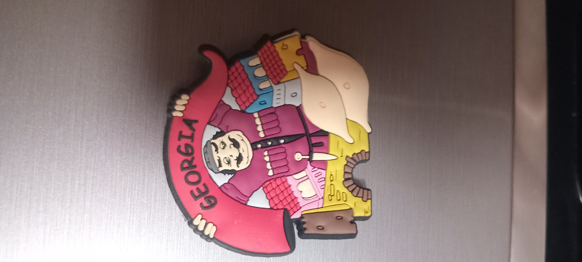 This is a close-up, horizontally oriented photograph of a novelty magnet on a metallic surface, likely a refrigerator. The magnet is colorful and depicts a cartoonish scene with a person and buildings.
The central subject is a male figure with a prominent mustache, wearing a traditional-looking, ornate maroon jacket with decorative trim and buttons. He is shown from the chest up, with his head tilted slightly. He appears to be holding a large, curved red banner with the word "GEORGIA" written in white, stylized block letters along its curve. The banner wraps around him, with his hands visible at either end, appearing to be made of a flexible material like rubber or plastic.
Behind the figure, a collection of stylized buildings is depicted. These structures have brightly colored roofs in shades of red and blue, and yellow and white walls. Some of the buildings have small windows indicated by black outlines and some have small circular openings. The architectural style is simplified and somewhat abstract, suggesting a whimsical representation of a town or village. To the right of the figure, there are two large, light-tan, bulbous shapes that could represent stylized trees, clouds, or even traditional Georgian hats (like the Papakhi).
The background of the image is a brushed metallic surface, exhibiting subtle vertical lines. The lighting is even, with no distinct shadows, and the time of day and weather conditions are not discernible. The overall impression is that of a souvenir magnet, likely purchased as a memento of a visit to Georgia. There are no discernible emotions expressed by the figure, as it is a cartoon representation.