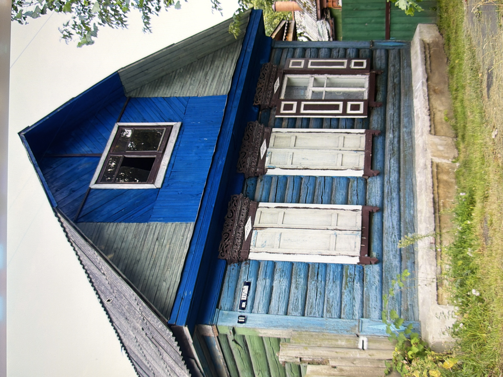 The image shows a close-up view of the front facade of a rustic, two-story wooden house painted in alternating horizontal bands of bright blue and faded green. The house has a steeply pitched roof covered with weathered grey corrugated metal. The upper story features a single, small window with a dark brown frame and a broken pane, set against the bright blue siding. The lower story has two windows with white shutters, and one window with dark brown shutters surrounding a white-framed pane. Both lower story windows are adorned with intricately carved wooden trim above them. A street sign with Cyrillic lettering and the number "11" is attached to the blue siding to the left of the left window. The foundation of the house appears to be made of concrete, and there is a low, concrete step leading up to the entrance. Lush green grass and some wildflowers grow in front of the house, with a wooden fence visible on the left side and a green metal fence on the right. In the background, glimpses of other houses and trees are visible, suggesting a residential area. The overall impression is of an old, somewhat dilapidated but charming dwelling in a rural or semi-rural setting, possibly within the city of Kyiv, Ukraine.