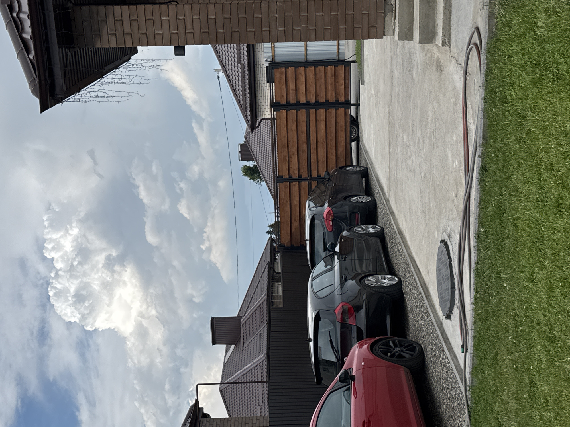 The image depicts a residential outdoor scene. In the foreground, a neatly mowed green lawn is visible, bordering a concrete pathway. A coiled garden hose lies on the edge of the concrete. Further back, several cars are parked on a gravel surface. These include a red car on the left, and two black cars parked side-by-side. Behind the cars, a dark wooden fence runs horizontally, with a modern wooden gate in the center. The background features residential buildings with brown tiled roofs and brick structures. The sky above is partly cloudy with a large, dramatic white cumulus cloud formation. The scene appears to be a driveway or yard area of a private residence. The location is specified as Дёмино, Russia.