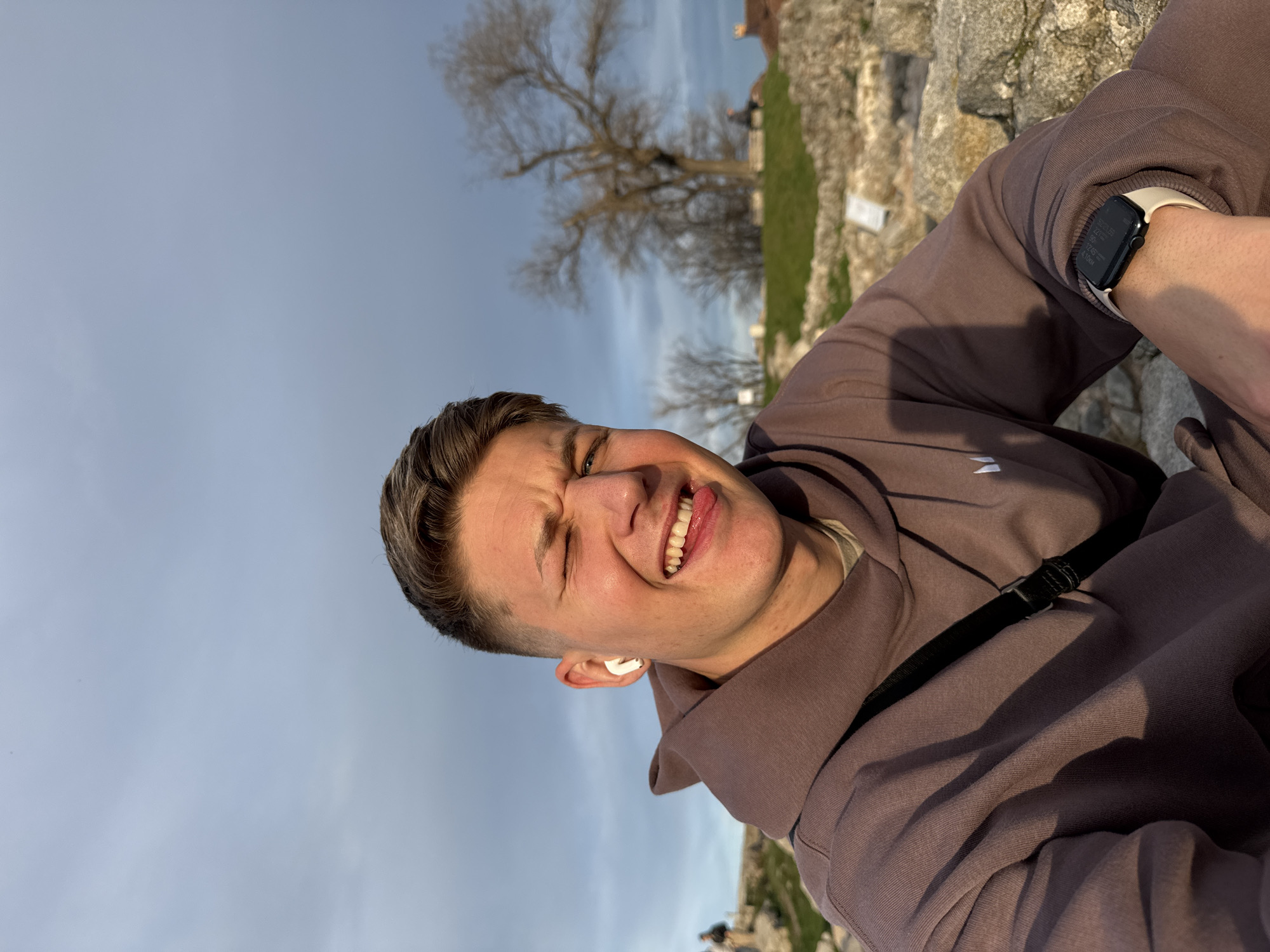 A portrait of a young man taking a selfie in Plovdiv, Bulgaria. He is light-skinned with short, light-brown hair and is wearing a mauve-colored hooded sweatshirt and a black shoulder bag strap. An Apple Watch is visible on his wrist. He is squinting and sticking out his tongue in a playful manner. In the background, there's a clear, slightly cloudy sky, the bare branches of a tree, and a stone structure, likely a part of the ancient ruins in Plovdiv. The image appears to be taken outdoors during the day.