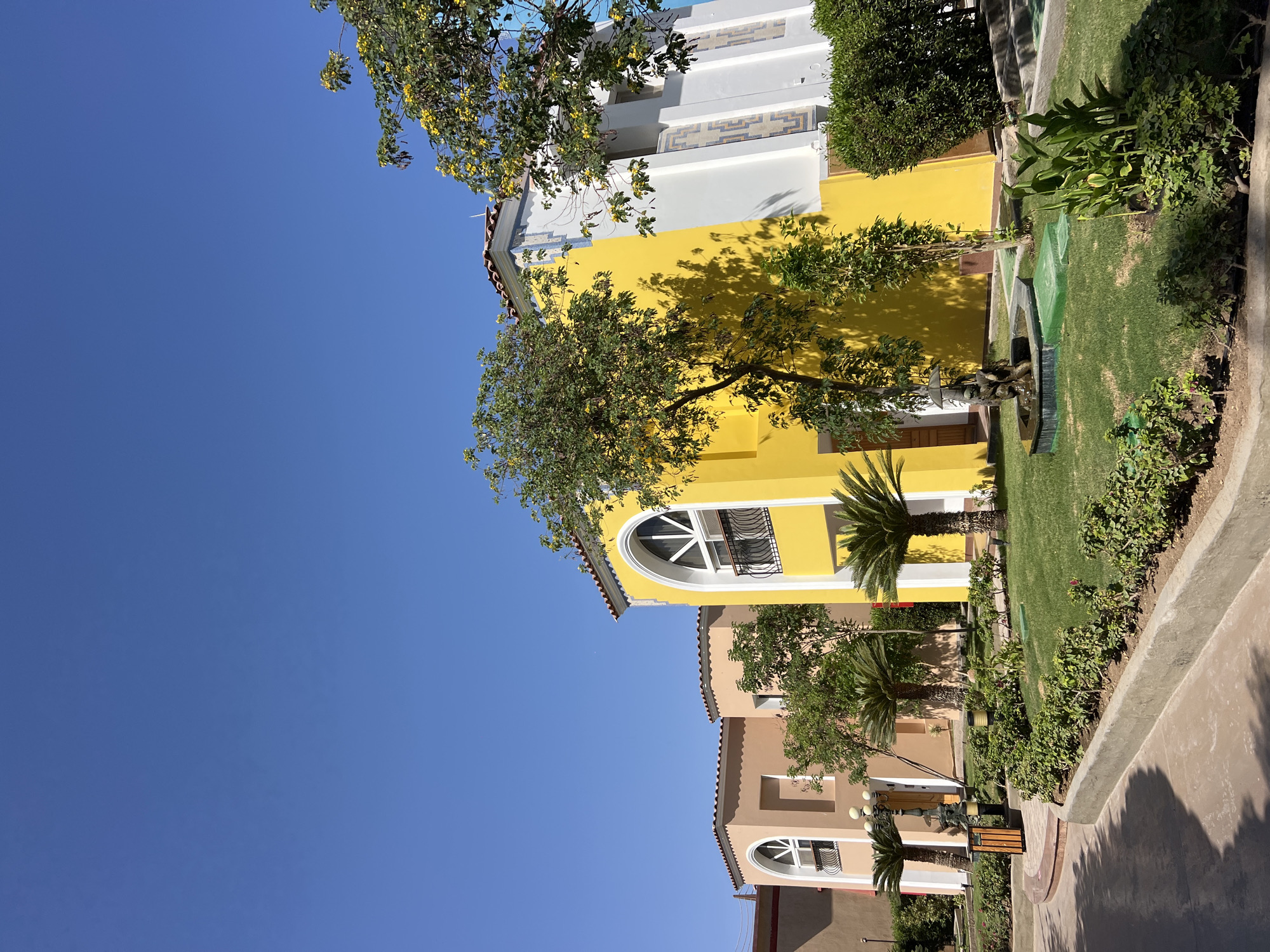The image depicts a row of Mediterranean-style buildings in Sharm El Sheikh, Egypt. The buildings are painted in warm hues of yellow, peach, and white, with red-tiled roofs and arched windows. Lush green landscaping, including palm trees and manicured bushes, surrounds the buildings. A clear blue sky dominates the upper portion of the frame. The scene appears tranquil and well-maintained, suggesting a resort or residential complex. There are no people visible in the image.
