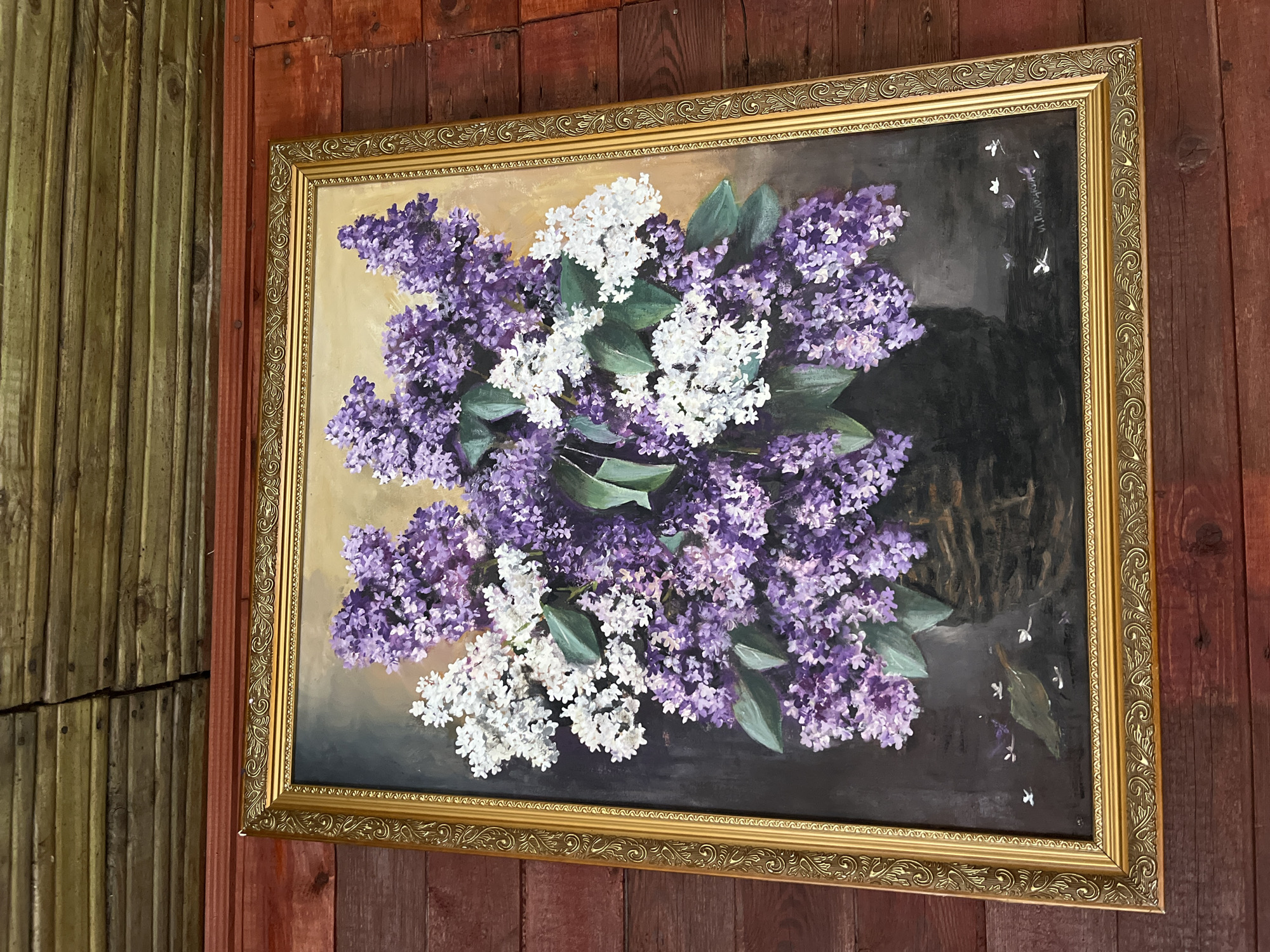The image shows an oil painting of a floral bouquet hanging on a wooden wall. The bouquet, dominated by purple and white lilacs, is arranged in a vase against a dark, somber background with subtle hints of light. The painting has an ornate gold frame. The wall appears to be constructed from vertical wooden planks, possibly tongue and groove style, in a reddish-brown tone. The location is Мшинская, Russia.