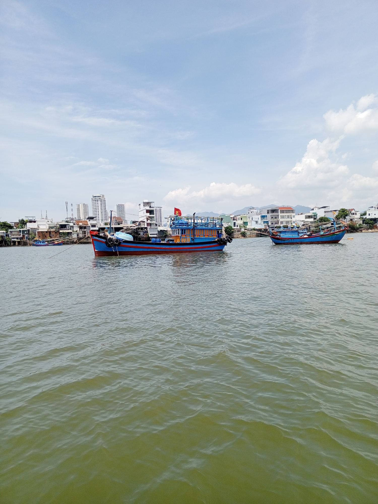 Two blue and red fishing boats are anchored in the water, with a city skyline in the background. The boat on the left is larger and more detailed, featuring a red hull with a blue stripe and several decks. It is tied to another, smaller blue boat in the distance. The boat on the right is smaller and simpler in design, also with a blue hull and red trim. Both boats appear to be in a calm body of water, likely a bay or river, given the presence of the cityscape. The background shows a cluster of buildings, ranging from low-rise structures to taller, modern high-rises. The sky is partly cloudy with a light blue hue. The overall scene depicts a typical day in a coastal city where fishing is a prominent activity.