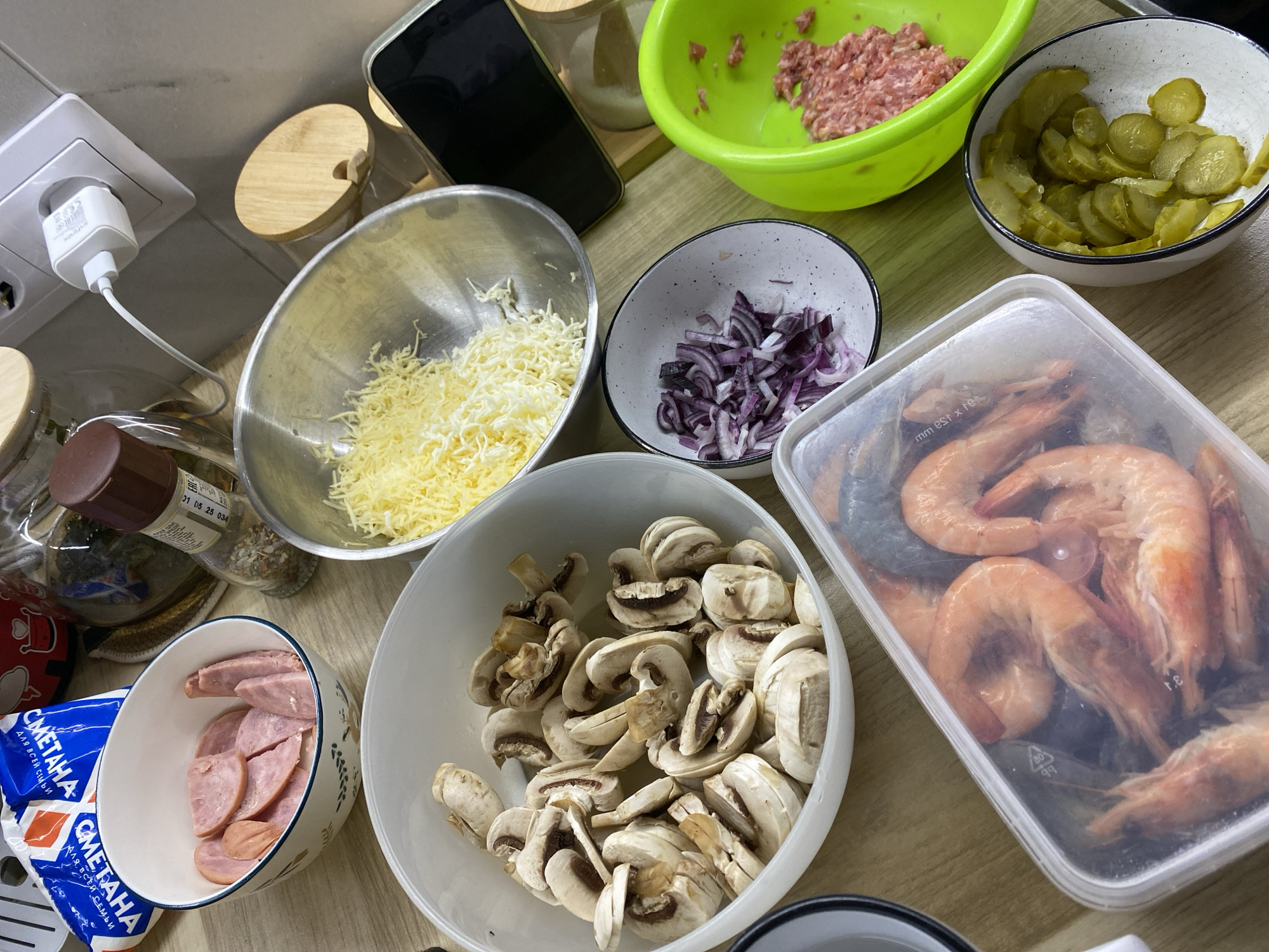 This indoor scene shows ingredients prepared for cooking. There are bowls containing sliced mushrooms, sliced red onion, shredded cheese, sliced sausage, cooked shrimp, sliced pickles, and ground meat. Jars and containers of various ingredients and seasonings surround the bowls. A charger is plugged into a wall outlet. A packet of Smetana (sour cream) for sale in Izhevsk, Russia is partially visible.