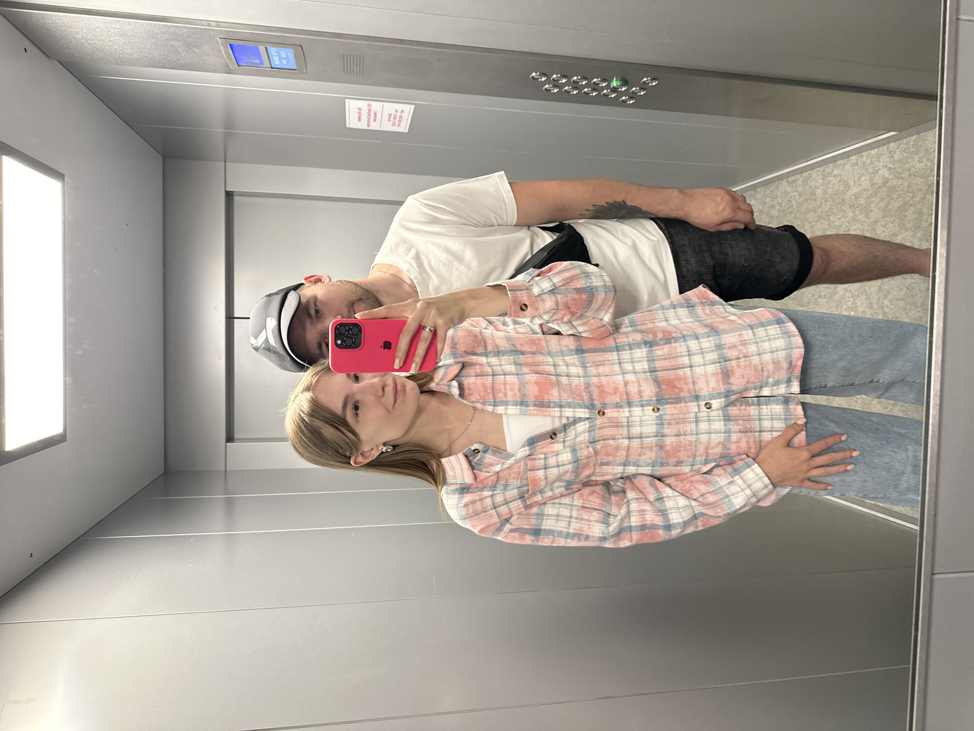 A young adult male and female are taking a selfie in an elevator. The male wears a white t-shirt, dark shorts, and a baseball cap. He has a tattoo on his upper arm. The female wears a pink and blue plaid shirt, jeans, and a white undershirt. The cell phone is pink. The elevator has metal walls and a panel with buttons and a digital display. A small sign is present. The location is likely a building in Chaikovsky, Russia.