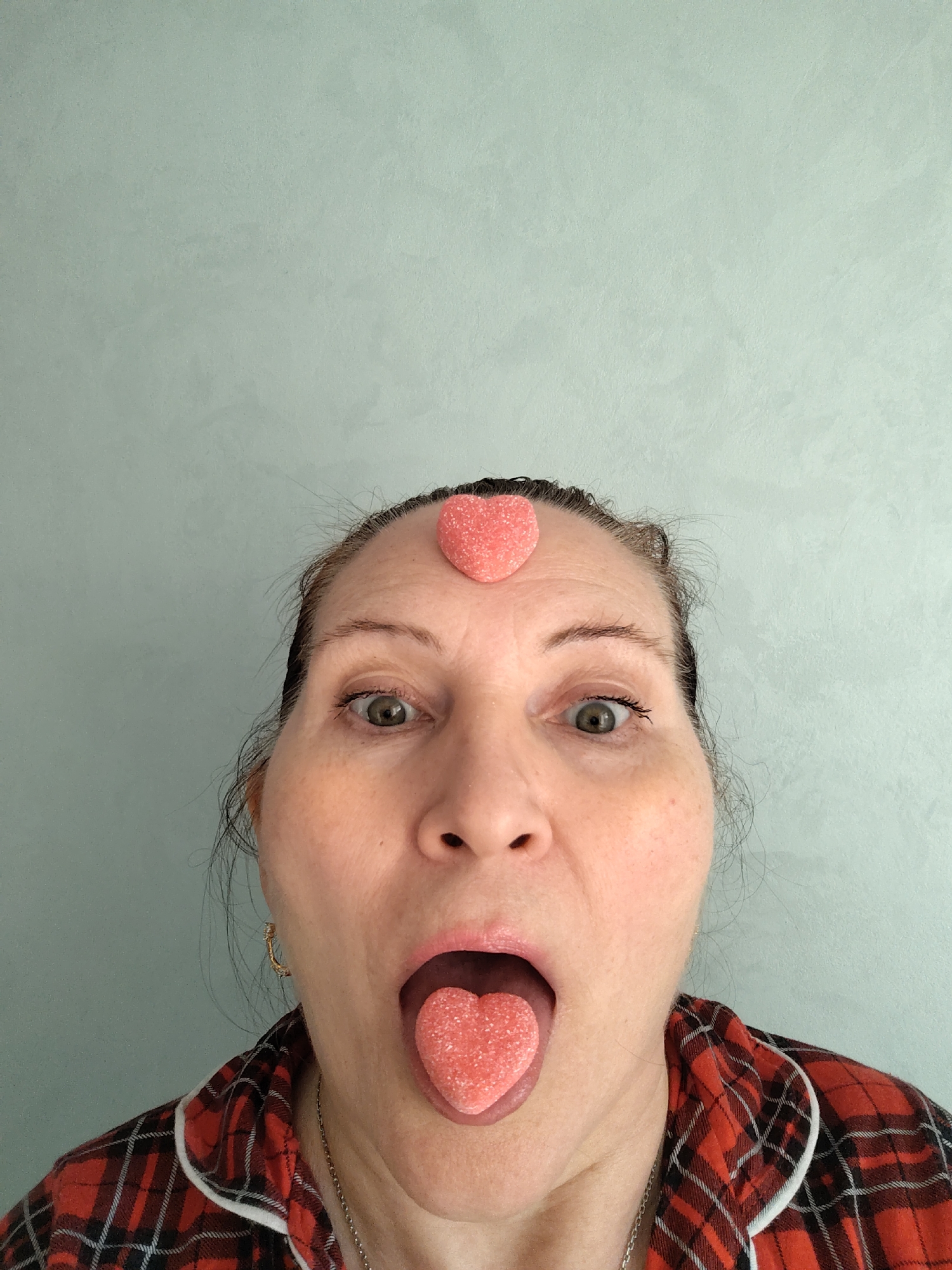 A woman with light skin and brown hair pulled back is centered against a light blue textured wall. She has a surprised expression, with wide eyes and her mouth open with her tongue sticking out. A heart-shaped candy is balanced on her forehead and another is on her tongue. She is wearing a red and black plaid shirt with a white trim, and a silver necklace. She also appears to be wearing silver earrings. The location of the image is not discernable.