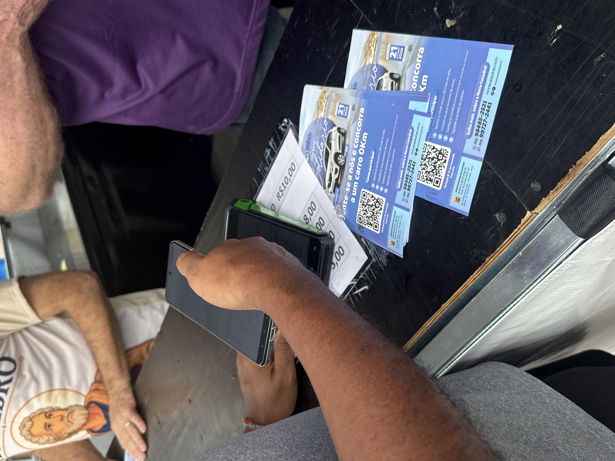 The image captures a close-up of a dark, wooden surface, likely a counter or table, with various objects placed upon it. In the foreground, a person's arms and hands are visible, with one hand holding a dark smartphone. Adjacent to the smartphone are two promotional flyers with a blue background, featuring images of a white car, a QR code, and text in Portuguese. One flyer contains the text "Junte-se a nós e concorra a um carro 0Km" which translates to "Join us and compete for a new car." Another flyer mentions "Sorteio Solidário" (Solidarity Draw) and "concorra km" (compete kilometers). Below the flyers, parts of what appear to be tickets or receipts are visible, with the numbers "8,00" and "5,00" printed on them, as well as "R$10,00". In the background, a person wearing a light-colored t-shirt with a graphic of a bearded man in a halo is partially visible, along with another person wearing a vibrant purple shirt. The promotional materials include information such as phone numbers, a website (novacatedral.com), and QR codes, indicating a promotional event or charitable campaign. One flyer explicitly mentions "Paróquia N° 5ª de Assunção Cabo Frio" which translates to "Parish of the 5th Assumption Cabo Frio," indicating that the event is associated with this parish in the city of Cabo Frio, Brazil.