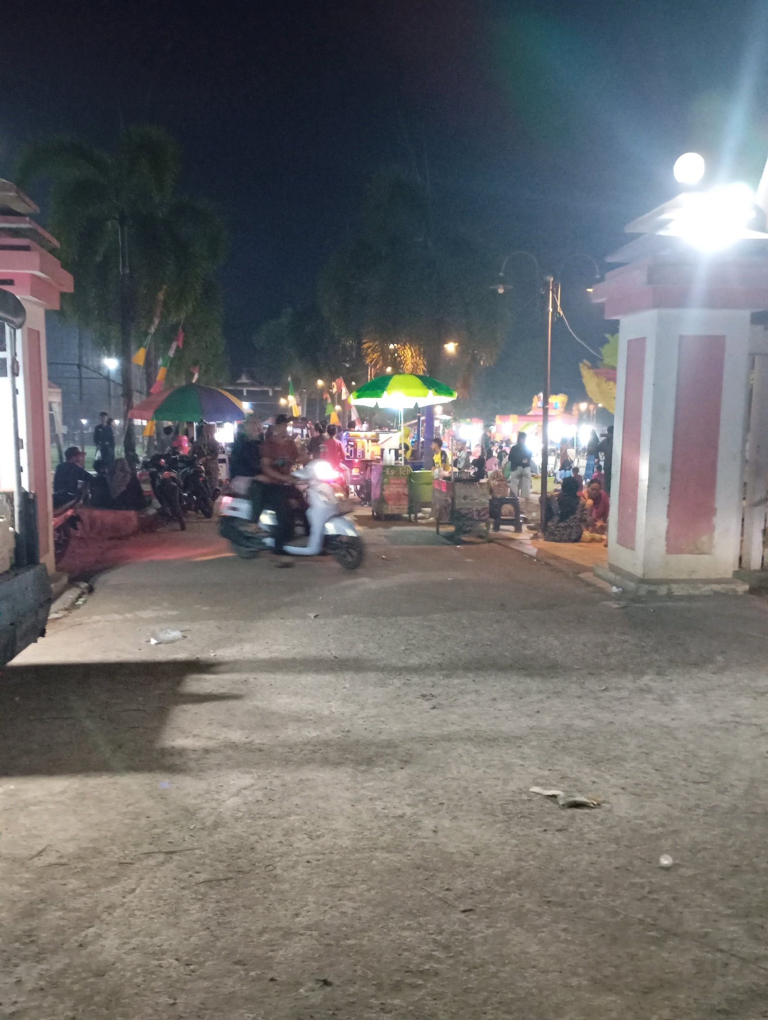 In the city of Kecamatan Haurgeulis, Indonesia, at night, a bustling scene unfolds at what appears to be an outdoor market or fair. In the foreground, a paved area is visible with some scattered debris. To the left, a portion of a building or structure with pink and white accents and a light fixture is prominent. Further into the scene, a group of people are gathered around stalls and vendors. Several motorcycles are parked, and some people are seated. In the middle ground, two individuals are riding on a white scooter. The background is filled with more activity, including people, food stalls with colorful umbrellas, and decorative lights, suggesting a lively evening event. Trees and palm trees are also visible in the background, contributing to the outdoor setting. The overall atmosphere is illuminated by artificial lights, indicating it is after dark.