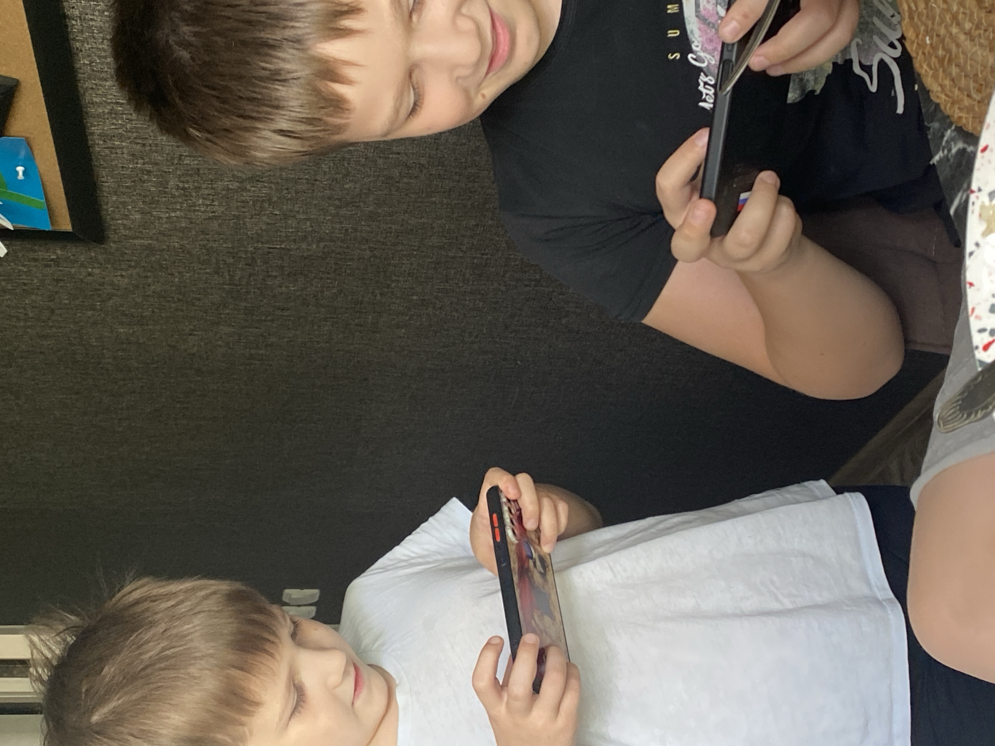 The image depicts two young boys engrossed in their mobile phones. The boy on the left, wearing a white t-shirt, is holding his phone horizontally, with his thumbs actively engaged on the screen. The phone has a colorful case with what appears to be an anime-style illustration. The boy on the right, in a black t-shirt with a graphic print, is also holding his phone horizontally and appears to be playing a game or watching something. The background suggests an indoor setting, possibly a dining area, with a dark, textured wall and a corkboard with some colorful drawings on it visible in the upper right corner. The scene is likely taking place in a residential environment.