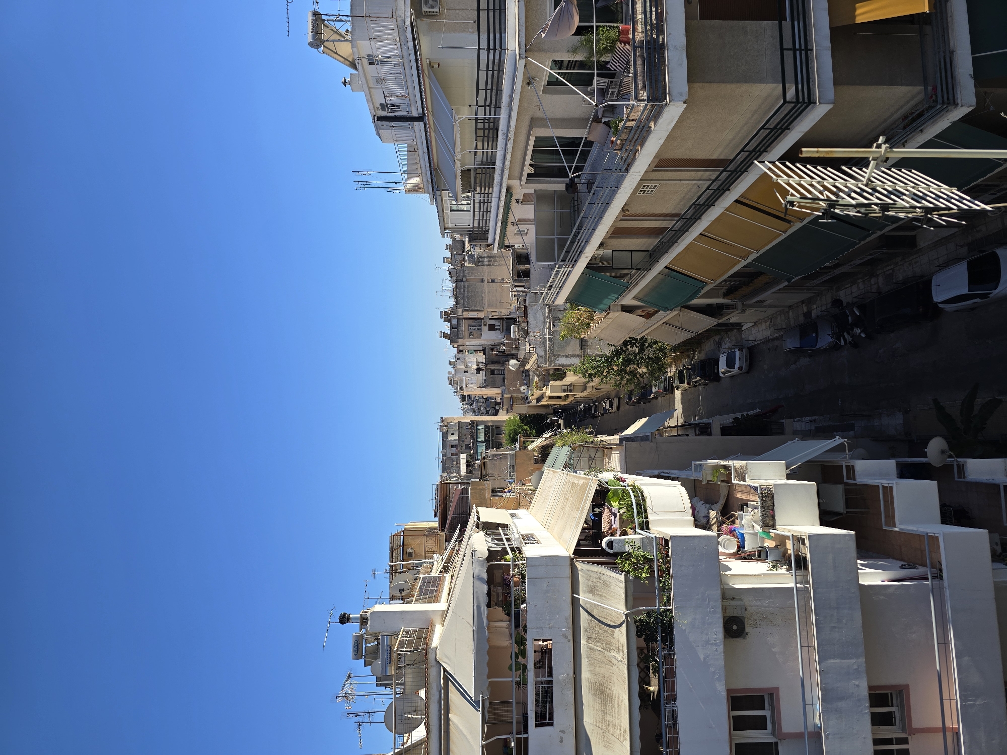 This is an outdoor, high-angle, full shot of a narrow city street in Athens, Greece, bordered by multi-story apartment buildings. The sky is clear and bright blue. The buildings on both sides of the street are densely packed, with balconies, awnings, and satellite dishes visible. Several cars and motorcycles are parked along the street. The architecture appears to be typical of residential areas in Athens, with a mix of older and more modern styles. There are no people clearly visible in the scene.