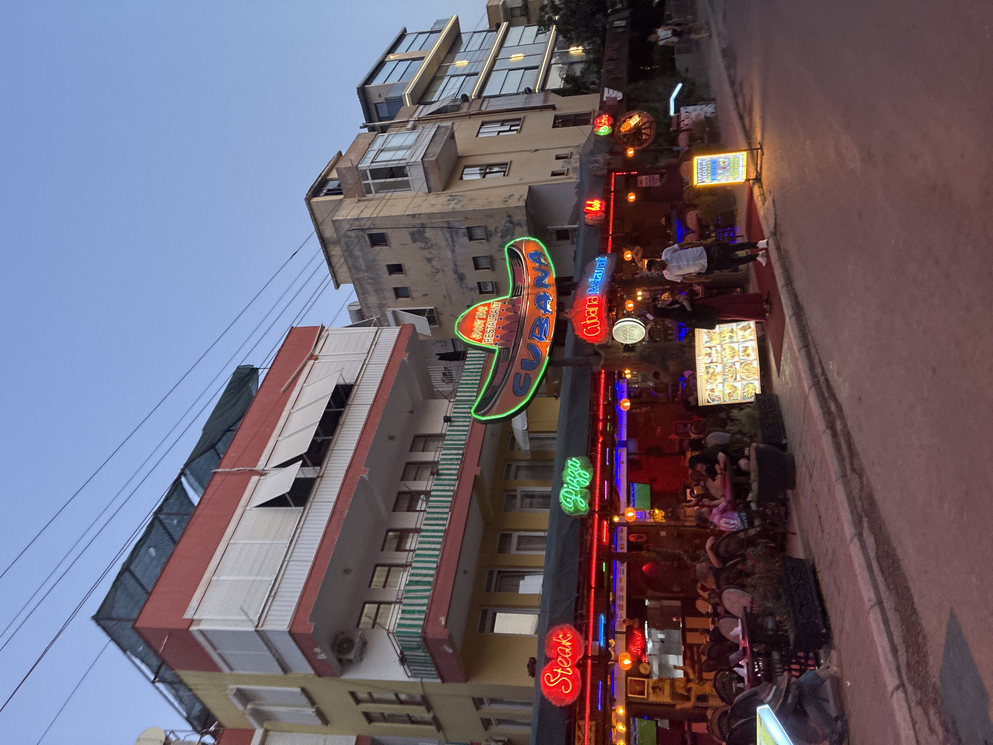 The image depicts a street scene in Alanya, Turkey, at dusk or early evening. The street is lined with buildings adorned with numerous neon signs, suggesting a lively entertainment district or restaurant area. Several people are visible, some seated at outdoor tables, indicating that businesses are open and patrons are dining or socializing.
Prominently featured is a large, colorful neon sign shaped like a sombrero, advertising "La MAMA" and "BOAT BAR RESTAURANT." Another neon sign displays "Cubana Restaurant." Other visible neon signs include "Pizza" and "Steak," along with a sign advertising "Happy Hour." A digital menu board displaying food images is also present.
The architecture suggests a mix of older and more modern buildings. Overhead wires are visible, a common sight in urban environments. The lighting from the neon signs creates a vibrant, festive atmosphere, contrasting with the fading natural light of the sky. The presence of outdoor seating and people engaged in activity implies a warm evening, suitable for al fresco dining. The overall impression is of a bustling nightlife scene in a tourist-oriented area.