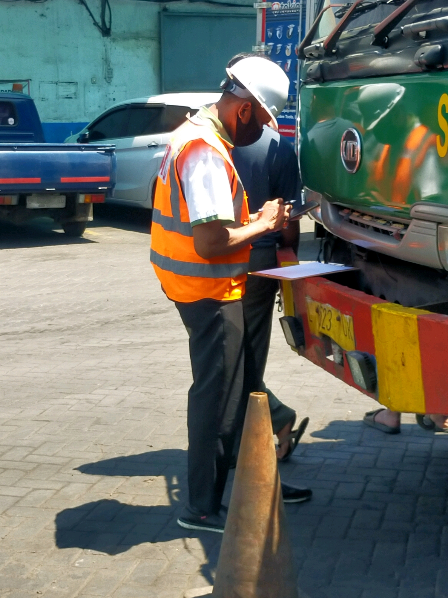 In a sunlit urban environment in Surabaya, Indonesia, a man wearing a white hard hat and an orange high-visibility vest over a dark shirt is writing on a clipboard. He is standing in front of a green truck, which has a red and yellow bumper. To the left of the man and the truck, a white car is parked. Further to the left, the rear of a blue pickup truck is visible. The ground is paved with interlocking bricks. A traffic cone is positioned in the foreground, to the right of the man. The scene appears to be an outdoor area, possibly a parking lot or a street.