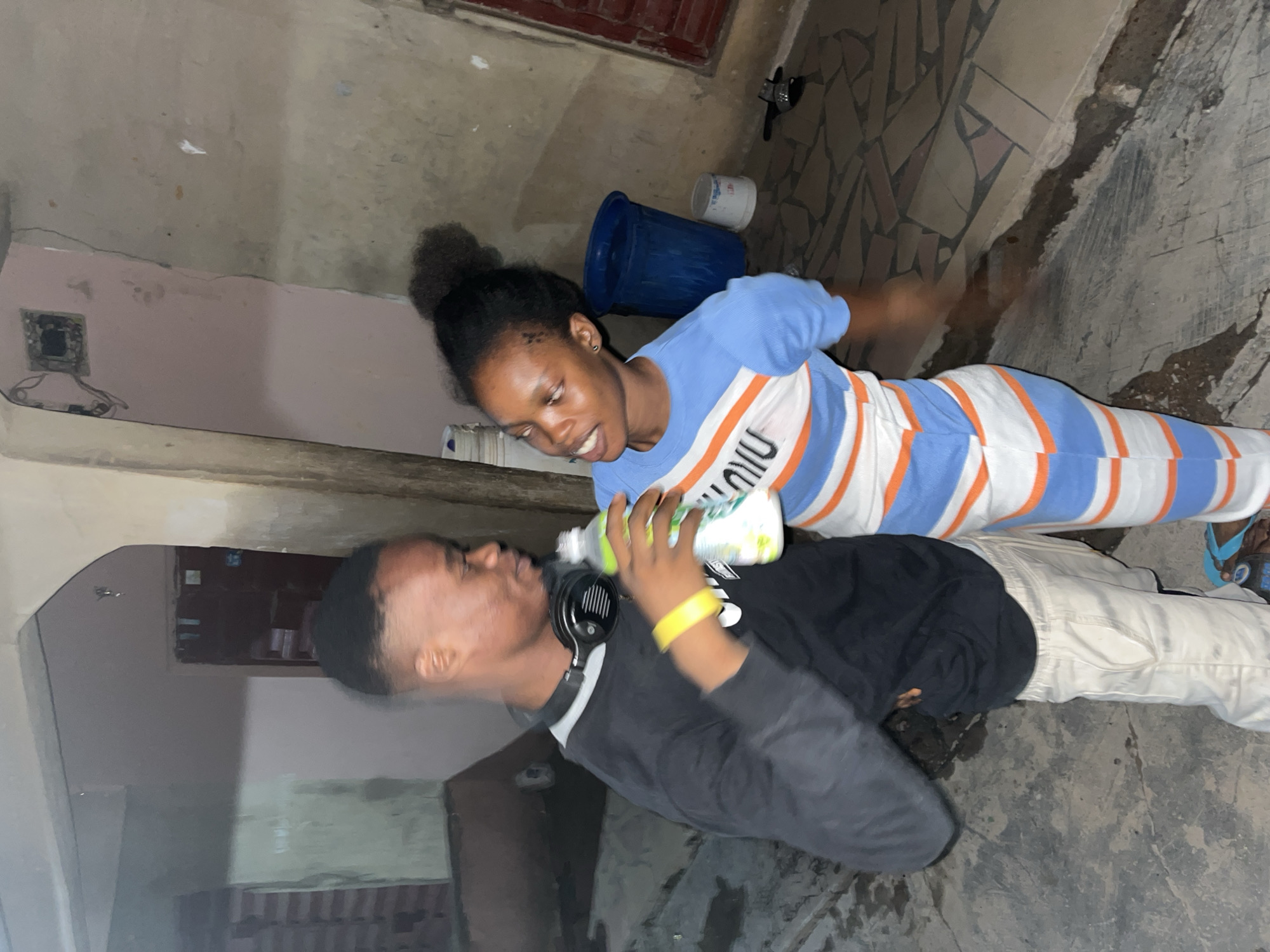 In an outdoor or semi-outdoor area in Unknown, Unknown, two young individuals are standing close together, engaging in a friendly interaction under artificial lighting.
On the left, a male, appearing to be in his late teens to early twenties, with dark skin and a fade haircut, wears a black long-sleeved shirt over a light undershirt, light-colored pants, and light blue flip-flops. Black over-ear headphones rest around his neck, and a yellow wristband is on his right wrist. He holds a clear plastic bottle containing a light green liquid in his right hand, looking towards the female with a slight smile.
On the right, a female, also appearing in her late teens to early twenties, with dark skin and dark hair tied in a top bun, wears a light blue short-sleeved top and matching pants/leggings with horizontal white and orange stripes. Black lettering, possibly "NIU," is visible within the white stripes of her top. She is looking at the male, smiling with her mouth slightly open. Her left hand is positioned near the bottle the male is holding, while her right arm shows motion blur.
The ground consists of various light-colored tiles and concrete with some darker, wet-looking patches. The background features textured, weathered walls of light brown or grey, an archway on the left, and visible cracks. Behind the female, a large dark blue plastic bucket, partially filled with water, and a smaller white bucket are present. An exposed electrical fixture with wires is visible on the upper right wall. A red door or shutter is seen on the far right, with a small metallic object resting on the ground nearby. The scene is illuminated, indicating evening or night.