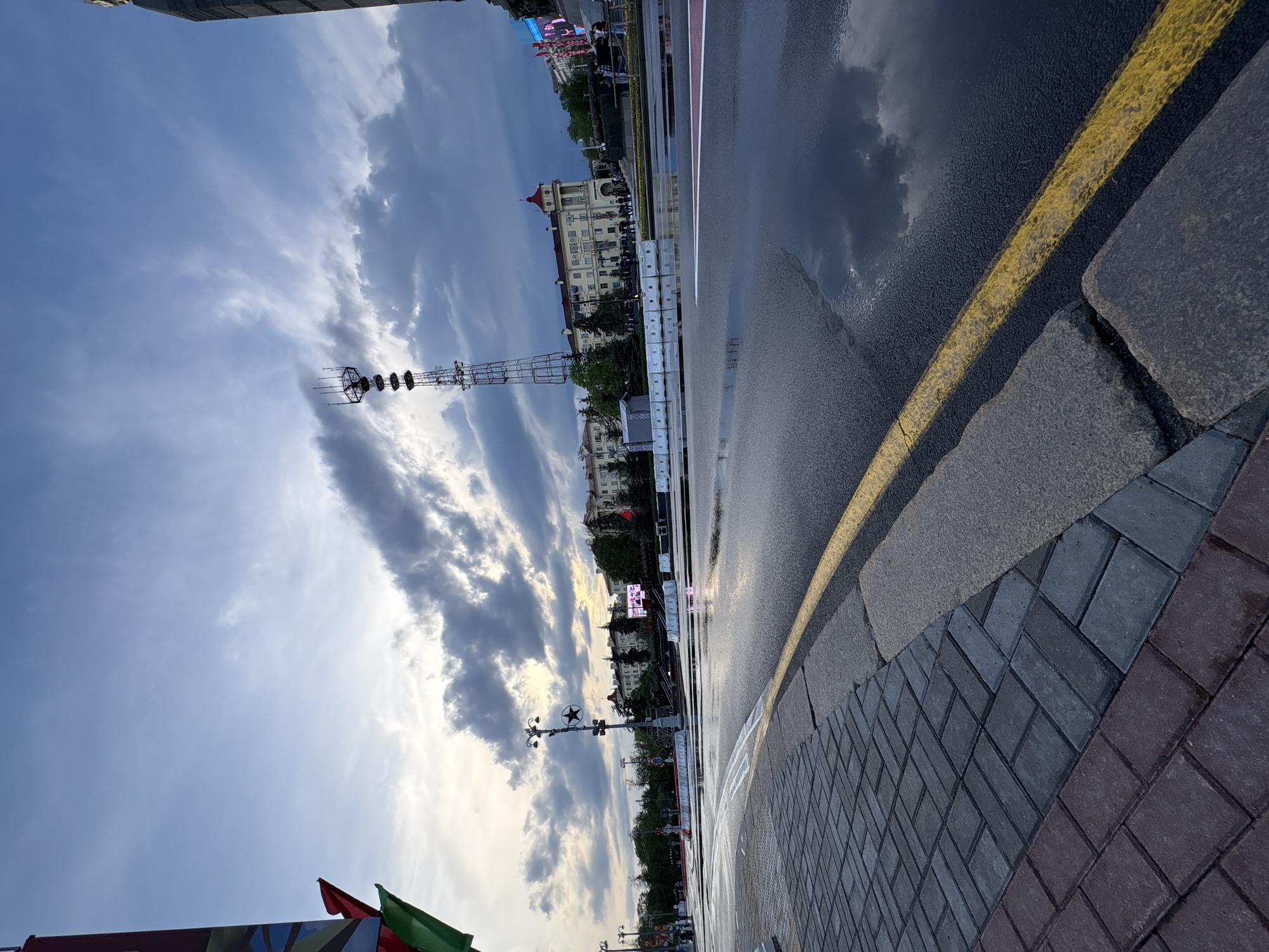 The image depicts a wet street scene in Minsk, Belarus, with reflections of the cloudy sky and buildings on the wet pavement. On the left is a brick sidewalk and a curb with a yellow line bordering the road. Across the street are buildings, trees, and streetlights. A communications tower stands prominently against the sky. People are seen walking on the sidewalks across the street. The image is taken from a low angle, emphasizing the texture of the pavement and the wet conditions.