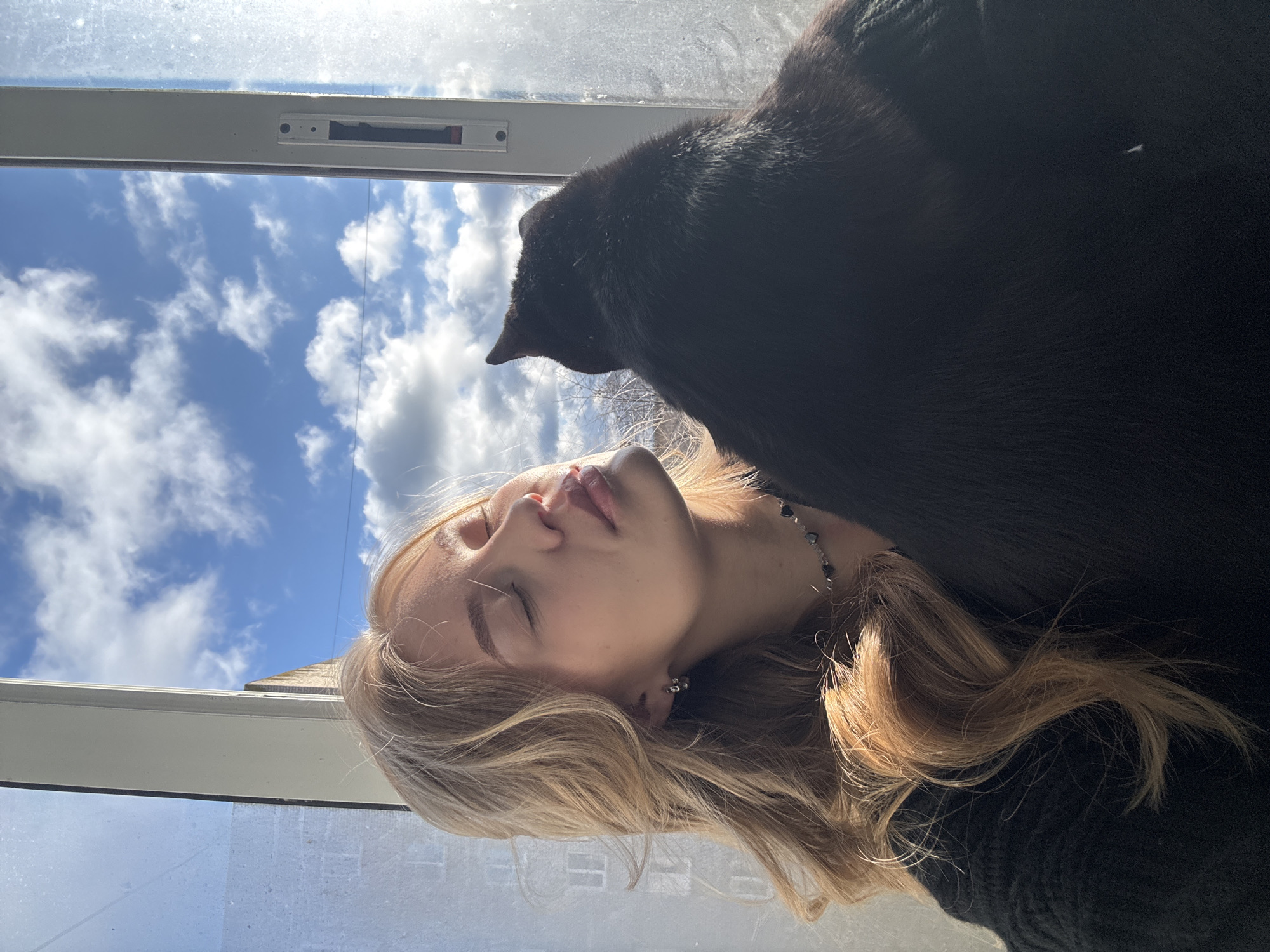 A serene portrait captures a young woman and a black cat relaxing by a window on a bright, sunny day in Chaikovsky, Russia. The woman, with long blonde hair, is lying down with her eyes closed, her face gently illuminated by natural light streaming through the window. She appears to be at peace, possibly resting or meditating. A small, dark bead necklace is visible around her neck, and she wears a simple silver earring.
Adjacent to her, a sleek black cat is also lying down, its back facing the viewer, positioned close to the woman's head. The cat's gaze or focus seems to be directed out the window, or it is simply resting alongside its companion.
Beyond the slightly smudged windowpane, a vibrant blue sky is visible, dotted with soft, white cumulus clouds, indicating clear and pleasant weather during the daytime. The white window frame is prominent, framing the outdoor view. The overall impression is one of quiet companionship and calm relaxation, bathed in natural light. No text is visible in the image.