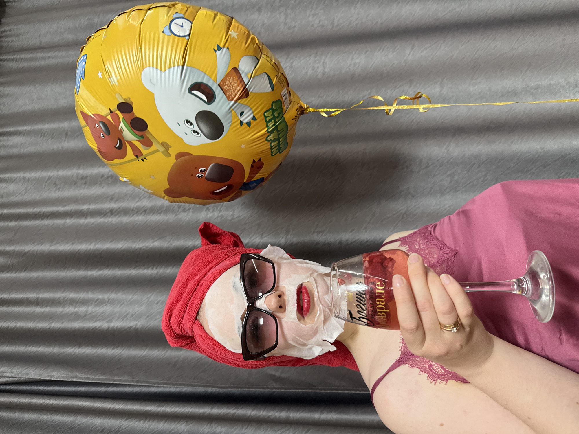 An indoor, medium shot captures a person lying down, enjoying a moment of relaxation or celebration. The image is rotated 90 degrees counter-clockwise, showing the person horizontally against a grey, subtly patterned background, possibly a bedspread or couch.
The main subject is a person, likely a woman, with a white sheet face mask applied to her face. She has a red towel wrapped around her head and is wearing dark-framed sunglasses over the mask. Her lips, visible through the mask's opening, are painted with red lipstick. She is wearing a pink or magenta top, possibly lingerie, with lace detailing. Her left hand, adorned with a ring, holds a stemmed glass containing a pink drink, possibly with raspberries or other fruit, suggesting a celebratory beverage.
Beside her, a large, yellow foil balloon floats, tethered by a golden ribbon. The balloon is decorated with cartoon bears and other small illustrations, including what appear to be alarm clocks, in a style reminiscent of "Masha and the Bear." Visible text on the balloon includes Russian phrases like "наш самый лучший" (our best). Text on the glass, also in Russian, reads "Пожелаю" (I wish you) and "в феврале" (in February), suggesting a wish or celebration connected to the month of February.
The overall scene conveys a mood of self-care and relaxation, possibly marking a personal celebration or indulgent moment.