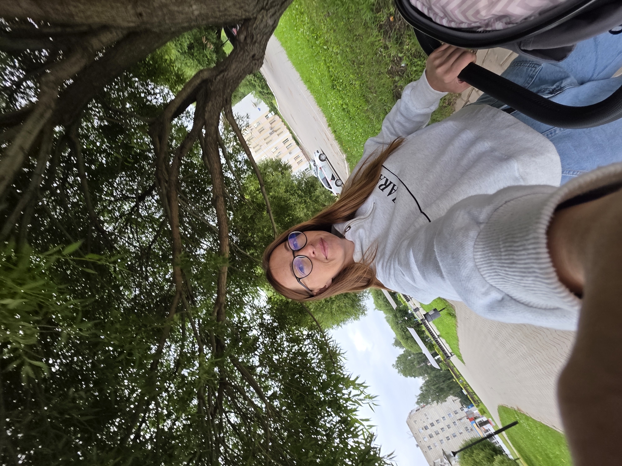 A woman with long brown hair and glasses is taking a selfie while pushing a stroller. She is wearing a light gray hooded sweatshirt with black lettering and blue jeans. She is standing on a paved walkway next to a grassy area. In the background, there are trees, a few buildings, and a road with cars. The environment appears to be a park or residential area in the city of Chaikovsky, Russia. The lighting suggests it is daytime, possibly overcast. The perspective of the photo is from the woman's point of view, holding the phone or camera.