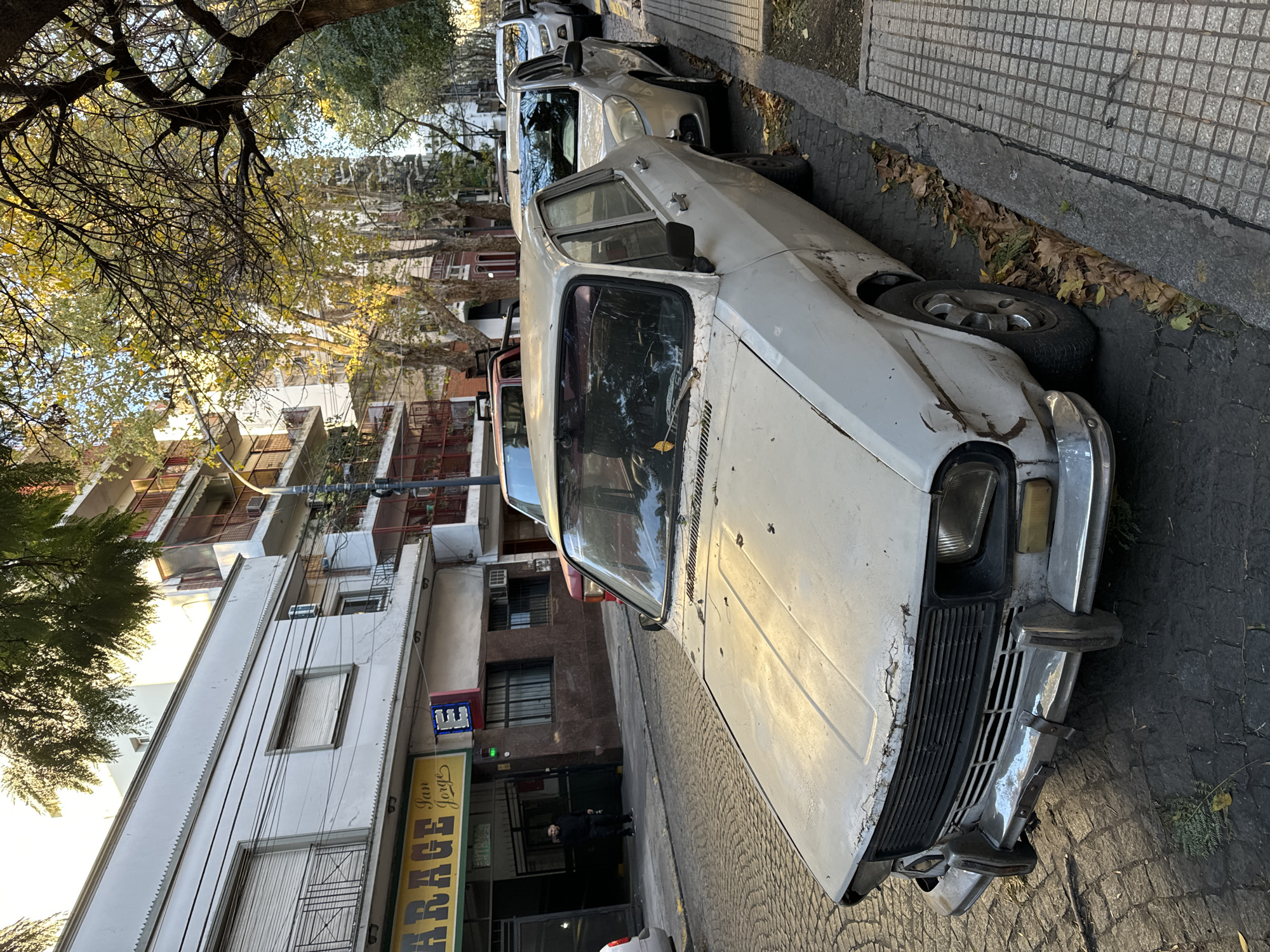 A white, older model car, possibly a Renault 12, is parked on a cobbled street in Buenos Aires, Argentina. The car shows signs of wear and tear, with damaged paint and a misaligned bumper. Behind it are other parked vehicles, including a light-colored modern car. On the left side of the image are buildings with balconies and shops. One shopfront is labeled "GARAGE." The trees lining the street indicate it is likely in a residential neighborhood. A person is standing near the entrance of the "GARAGE" shop. The environment appears to be an urban setting with autumn foliage on the trees and ground.