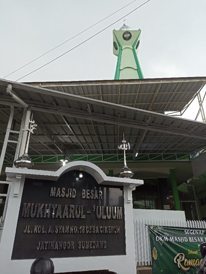 The image shows the facade of a mosque in Jatinangor, Indonesia. The mosque is called Masjid Besar Mukhtaarul-Ulum, located at Jl. Kola. Syam No. 18, Desa Cikeruh, Jatinangor, Sumedang. There is a sign with the mosque's name on it, and a minaret is visible in the background. The sign is a black rectangular sign with white letters. It is mounted on a white frame with a curved top. The minaret is tall and slender with a green and white color scheme. The building has a metal roof, and there is a green banner with white lettering to the right of the sign. The building also has a white-painted exterior with green accents. The sky is cloudy, and the image was taken during the day.