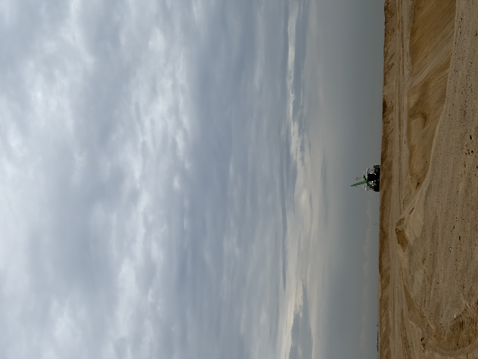 The image shows a sandy, barren landscape under a cloudy sky. In the distance, a green crane is visible on a vessel. The terrain appears to be a construction site or an area undergoing land reclamation, with mounds of sand and tire tracks on the ground. The sky is overcast with gray clouds. The scene suggests an industrial or developmental activity in an open, undeveloped area. There are no people visible in the image. The location in Alaguntan, Nigeria, is not discernible from the image itself, but the description is of the visual content.