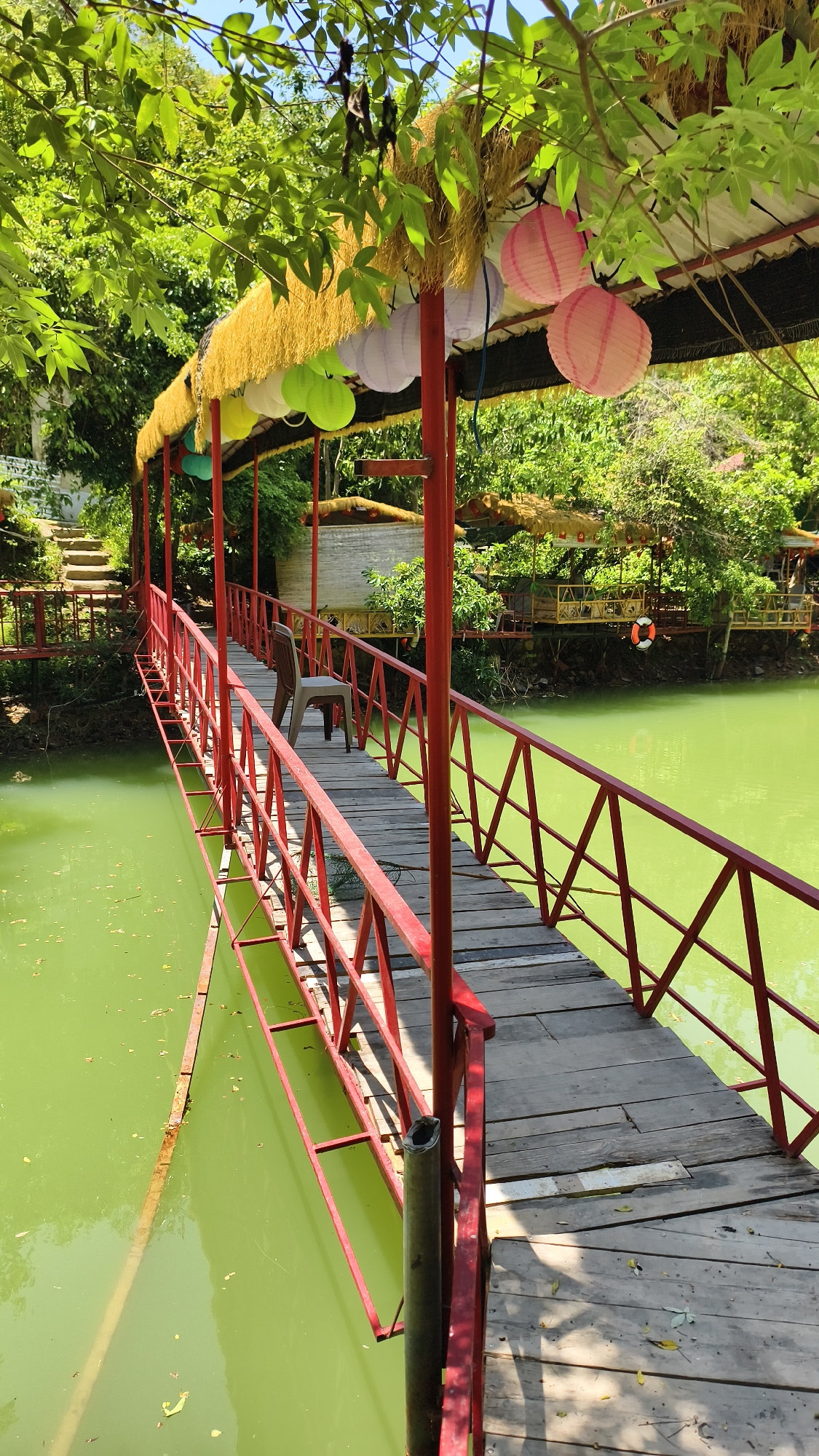 This is a medium shot, taken from a slightly elevated angle, of a wooden bridge with red metal railings that spans across a body of greenish water. The bridge has a thatched roof supported by red metal poles, and decorative paper lanterns in pink and light purple hang from the roof. A single brown plastic chair sits on the bridge, towards the far end. Trees with lush green foliage surround the scene, and the sky is visible overhead with a bright sun casting shadows on the bridge. In the background, there are several stilted structures with thatched roofs, suggesting a resort or restaurant setting. A life preserver ring is visible on one of the structures near the water's edge. The overall environment appears to be a serene, tropical outdoor location. Based on the context provided, this scene is located in Nam Nha Trang Ward, Vietnam.