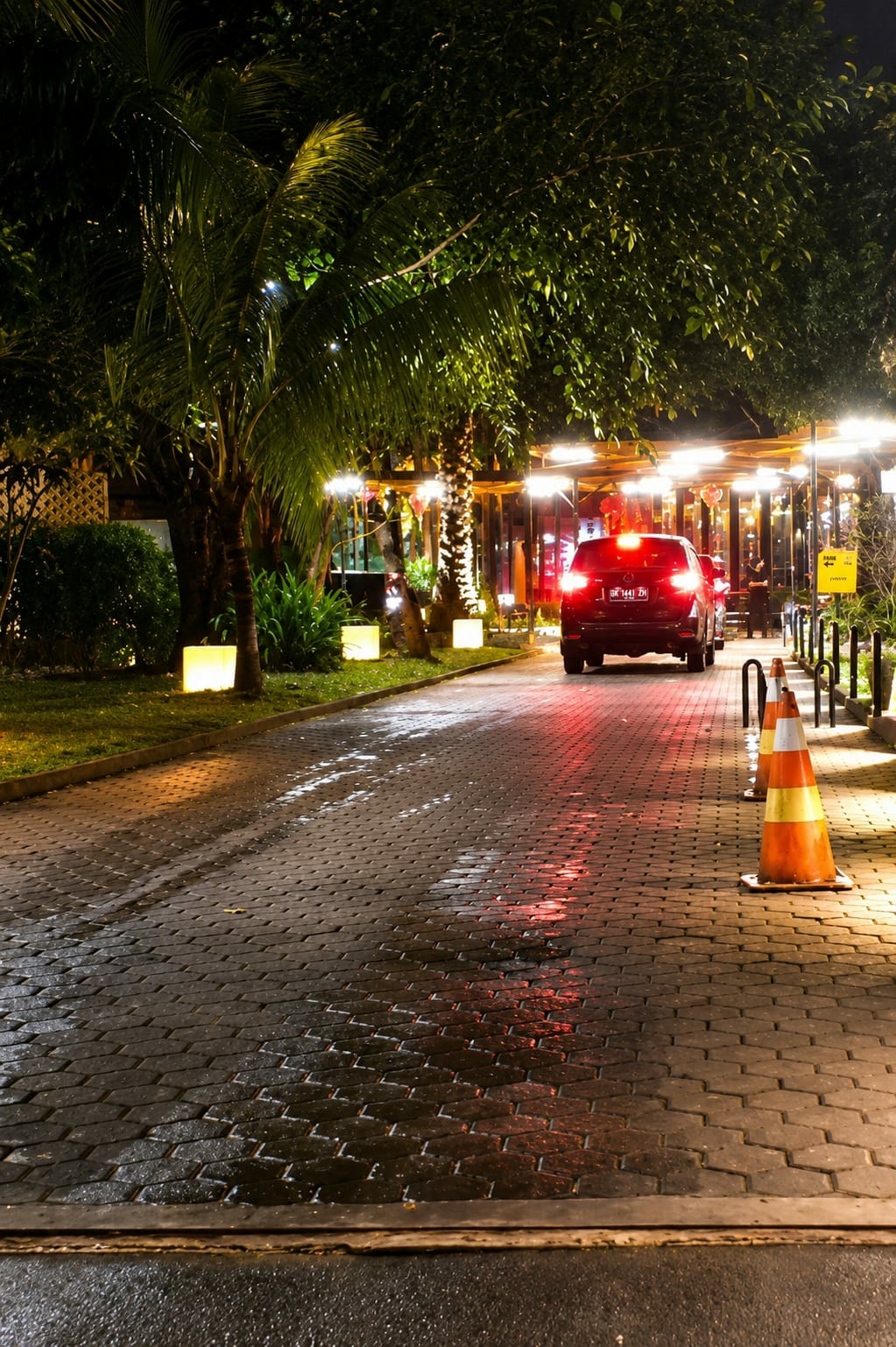 A dark-colored sedan with its brake lights illuminated is parked on a wet, cobblestone driveway at night. The car is positioned to the right of the frame, facing away from the viewer and towards a dimly lit establishment. The driveway is bordered by lush green grass and palm trees on the left, with strings of lights and foliage visible in the background. Several illuminated rectangular lamps are scattered across the lawn, casting a soft glow. Two orange traffic cones with yellow stripes are placed on the right side of the driveway, near the car. The overall scene suggests an entrance to a restaurant or commercial area in a tropical or subtropical urban setting, possibly City of Medan, Indonesia, given the tropical vegetation and architectural style.
