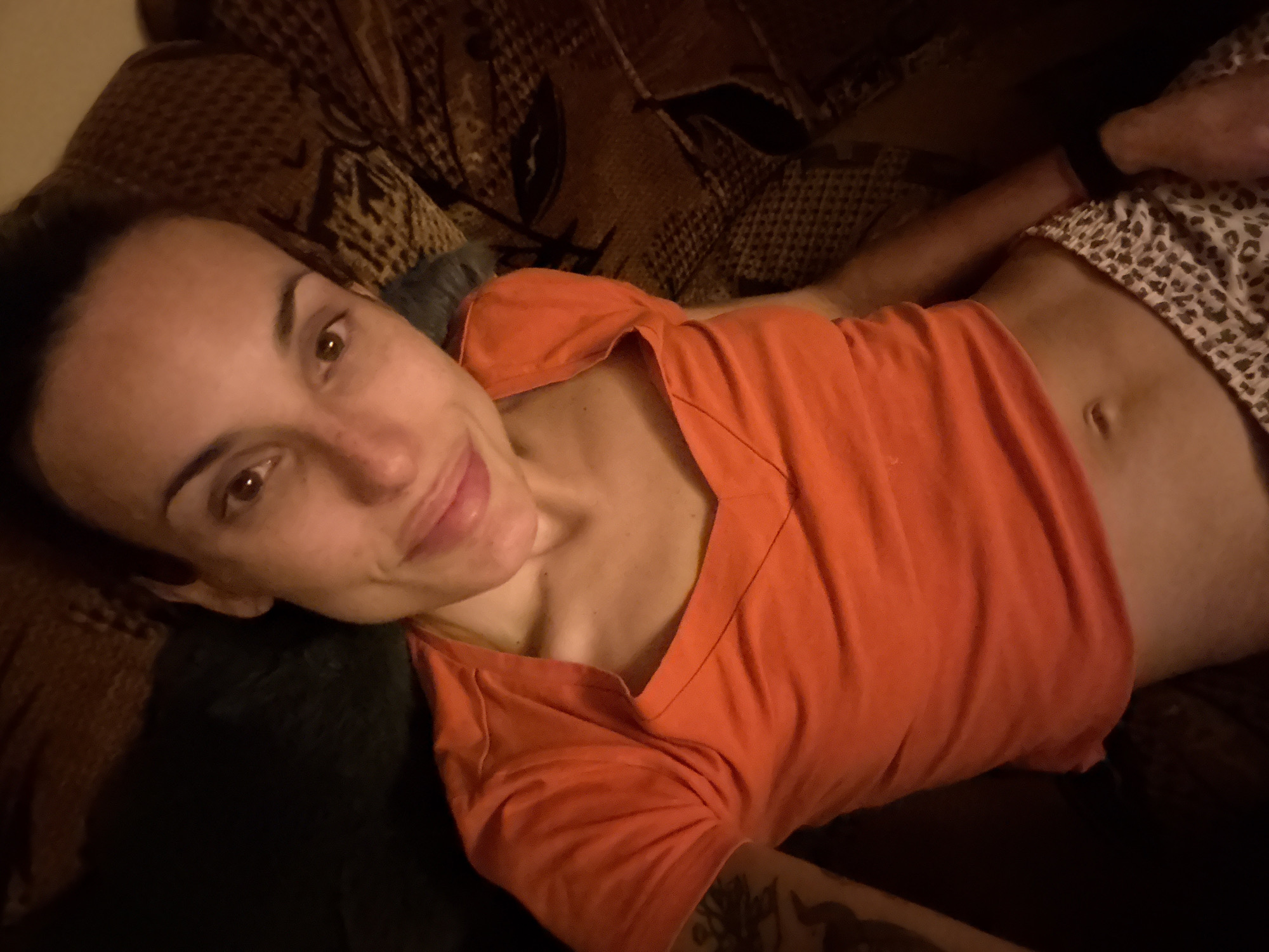A woman is depicted lying down indoors, looking directly at the camera with a relaxed and gentle smile. She has brown eyes, dark eyebrows, and slight freckles across her nose and cheeks. Her dark hair appears to be pulled back. She is wearing an orange, short-sleeved V-neck top and patterned shorts, possibly leopard print, which are visible around her midriff. A black watch or fitness tracker is on her right wrist, and a tattoo can be seen on her lower left arm.
The woman is resting on a surface covered with a brown and tan patterned fabric, which resembles an animal print or an abstract design, suggesting a couch, bed, or set of cushions. The lighting in the scene is dim and warm, indicating an indoor setting during the evening or night. The overall atmosphere appears calm and comfortable. No visible text is present in the image. The location context provided is Pleven, Bulgaria.